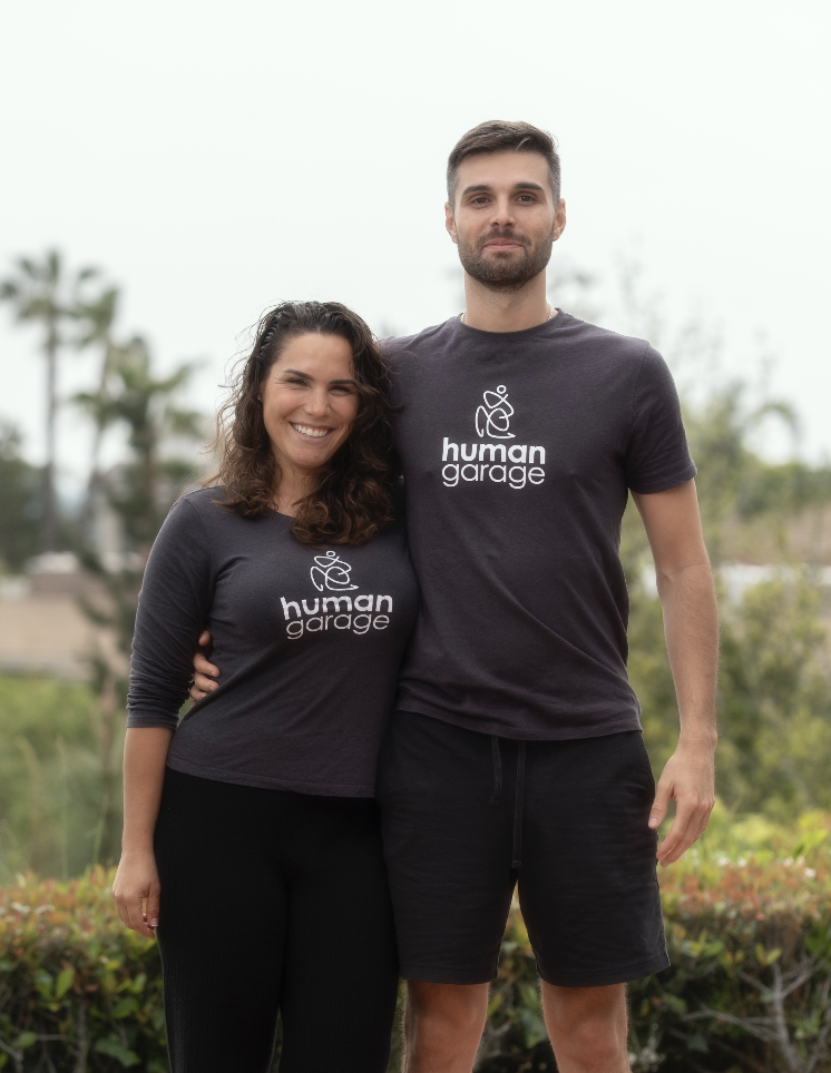 A man and woman standing outdoors, both wearing matching dark gray T-shirts with the logo 'human garage'. They are smiling and have their arms around each other, with a blurred background of trees and bushes.