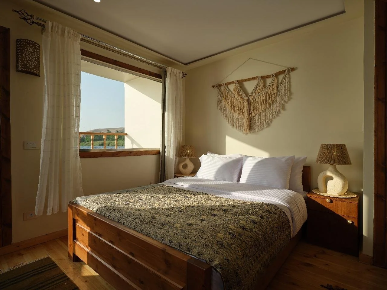Cozy bedroom with a wooden bed frame, patterned bedspread, white pillows, and natural light from a window with white curtains. Decor includes a macramé wall hanging and matching lamps on nightstands.