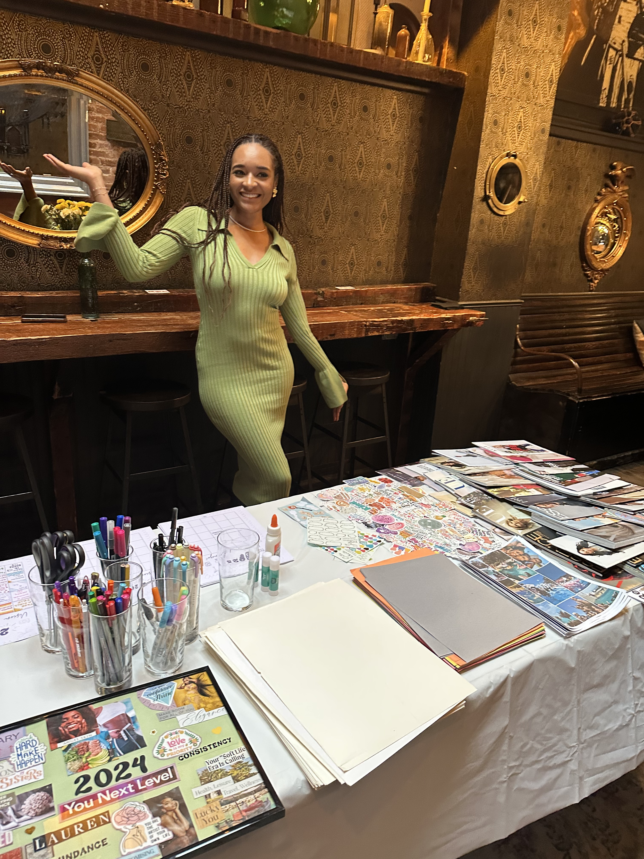 A woman in a long, green, ribbed dress standing at a table with various craft supplies and magazines in a warmly decorated room with gold framed mirrors and ornate décor.
