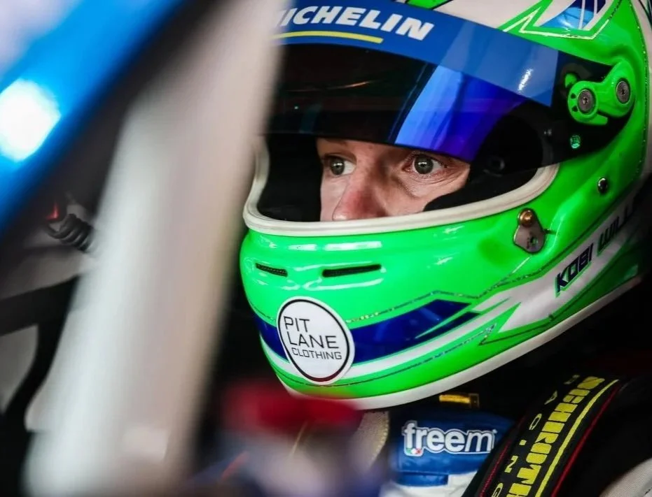 Close-up of a race car driver wearing a vibrant green helmet with blue and white accents, sitting in a race car. The helmet has a sticker that says 'PIT LANE CLOTHING'. The driver is focused, and part of the racing suit and interior of the car are visible.