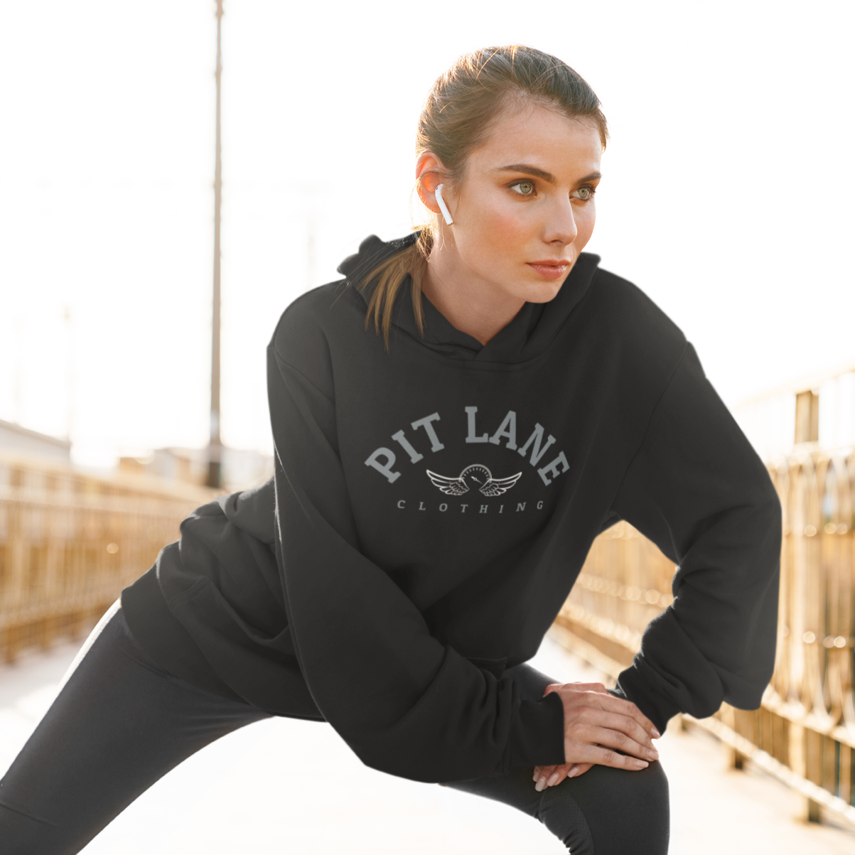 Person in sunglasses, black cap, hooded sweatshirt, and white pants kneeling on a racetrack.