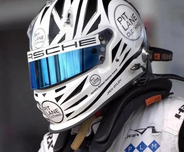 race car driver Jack Atley is seen wearing a white helmet with "Pit Lane Clothing" logo, inside a vehicle, focusing intensely through the visor. The helmet also has "Porsche" branding.