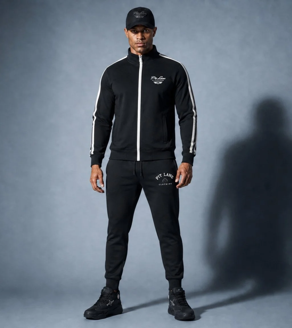 Man wearing black athletic tracksuit with white stripes, black cap, and black sneakers standing against a gray background.