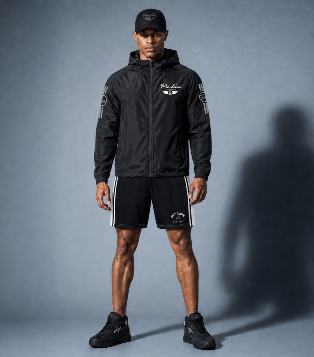 A man dressed in black athletic clothing, including a black cap, jacket, shorts, and sneakers, standing against a plain gray background.