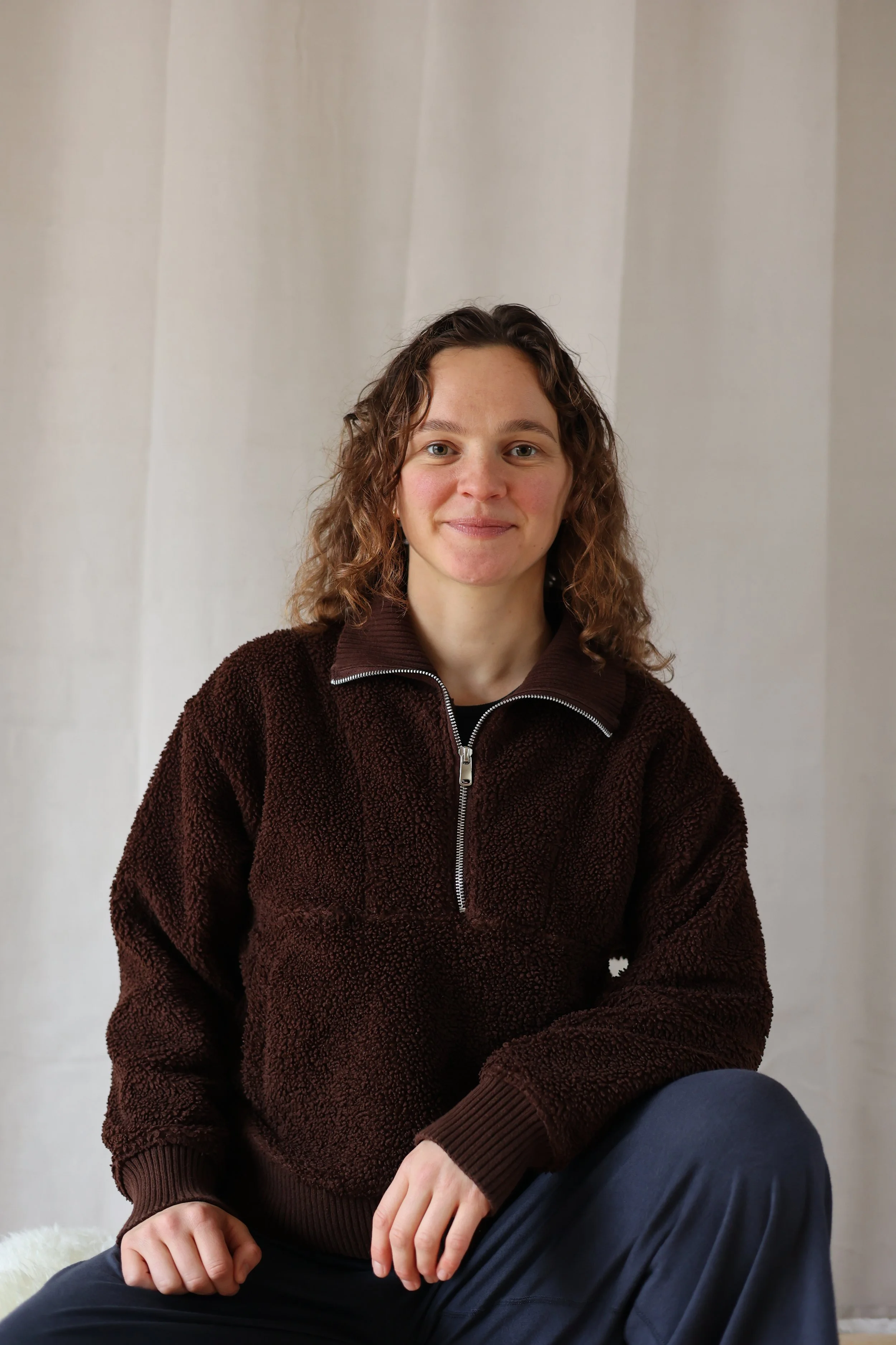 A woman taking a selfie in natural light, wearing a pink tank top and a gold necklace, with a plain white wall background.