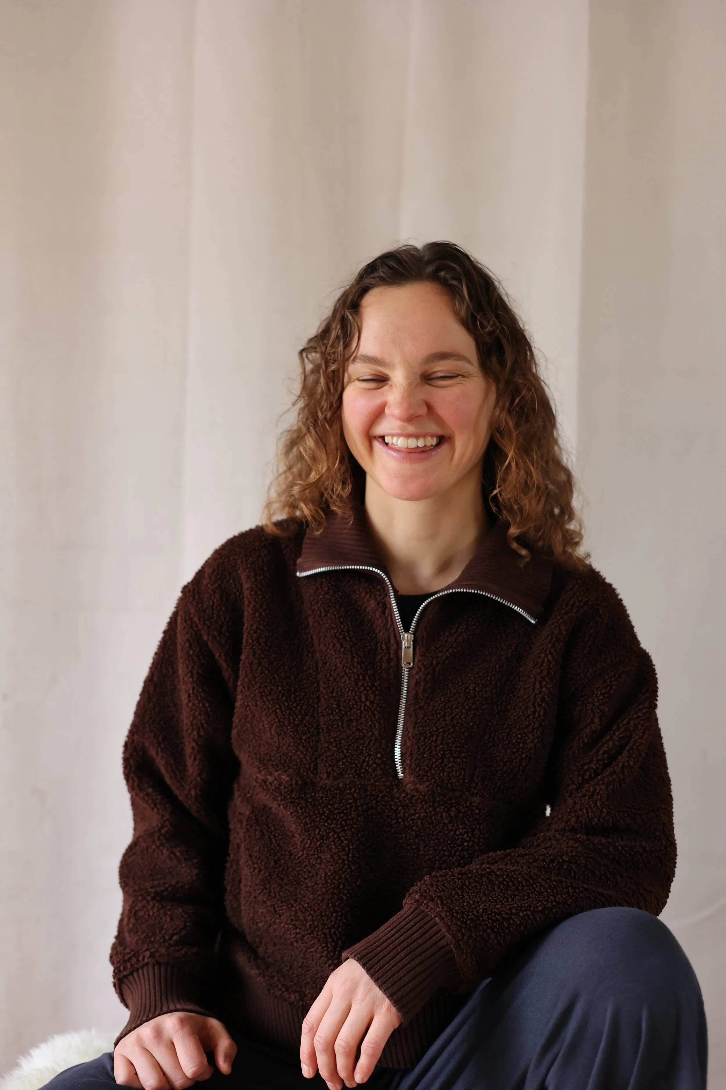 A woman taking a selfie in natural light, wearing a pink tank top and a gold necklace, with a plain white wall background.