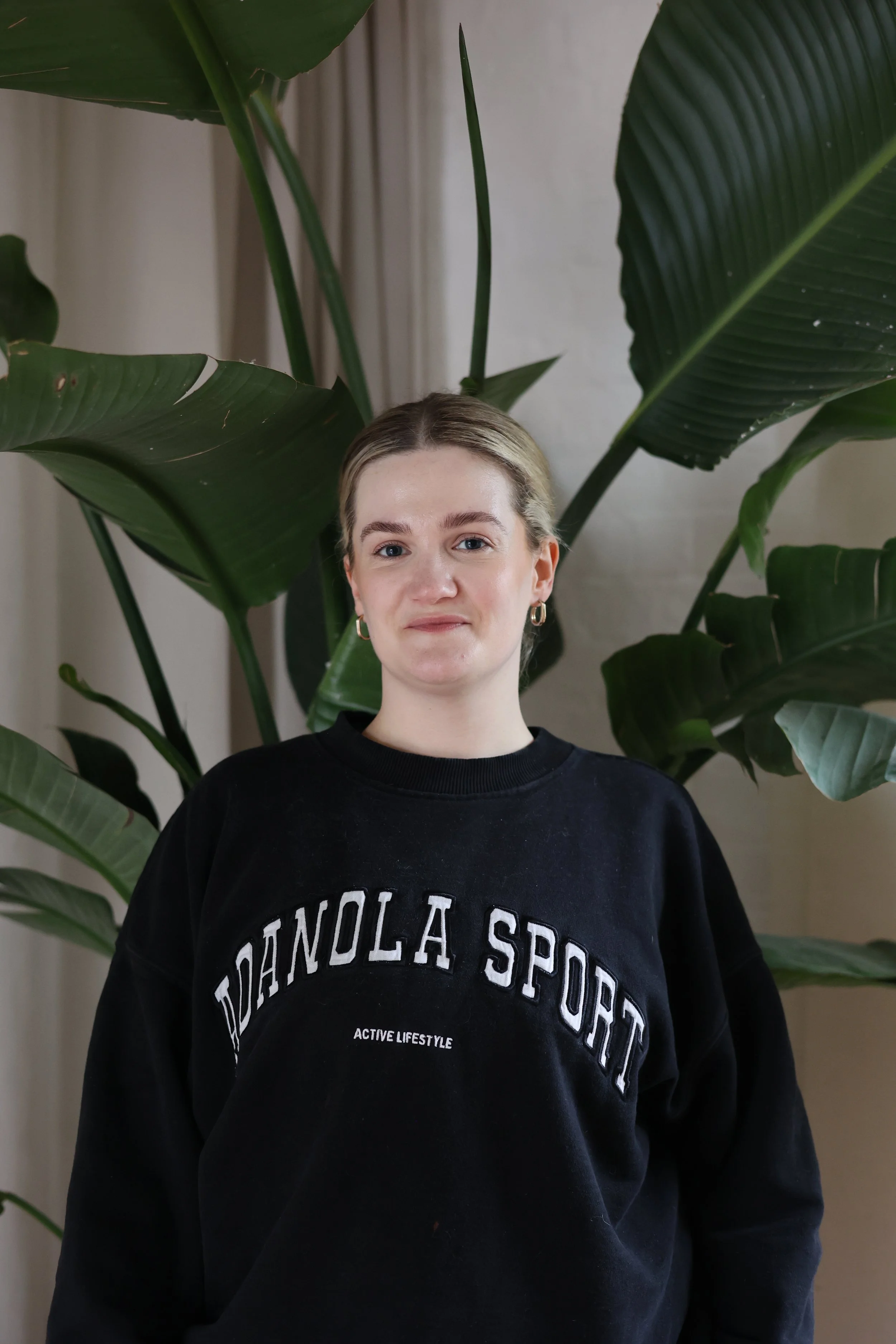 A woman with blonde, wavy hair wearing a black sleeveless top, standing against a light-colored plain wall, smiling softly.