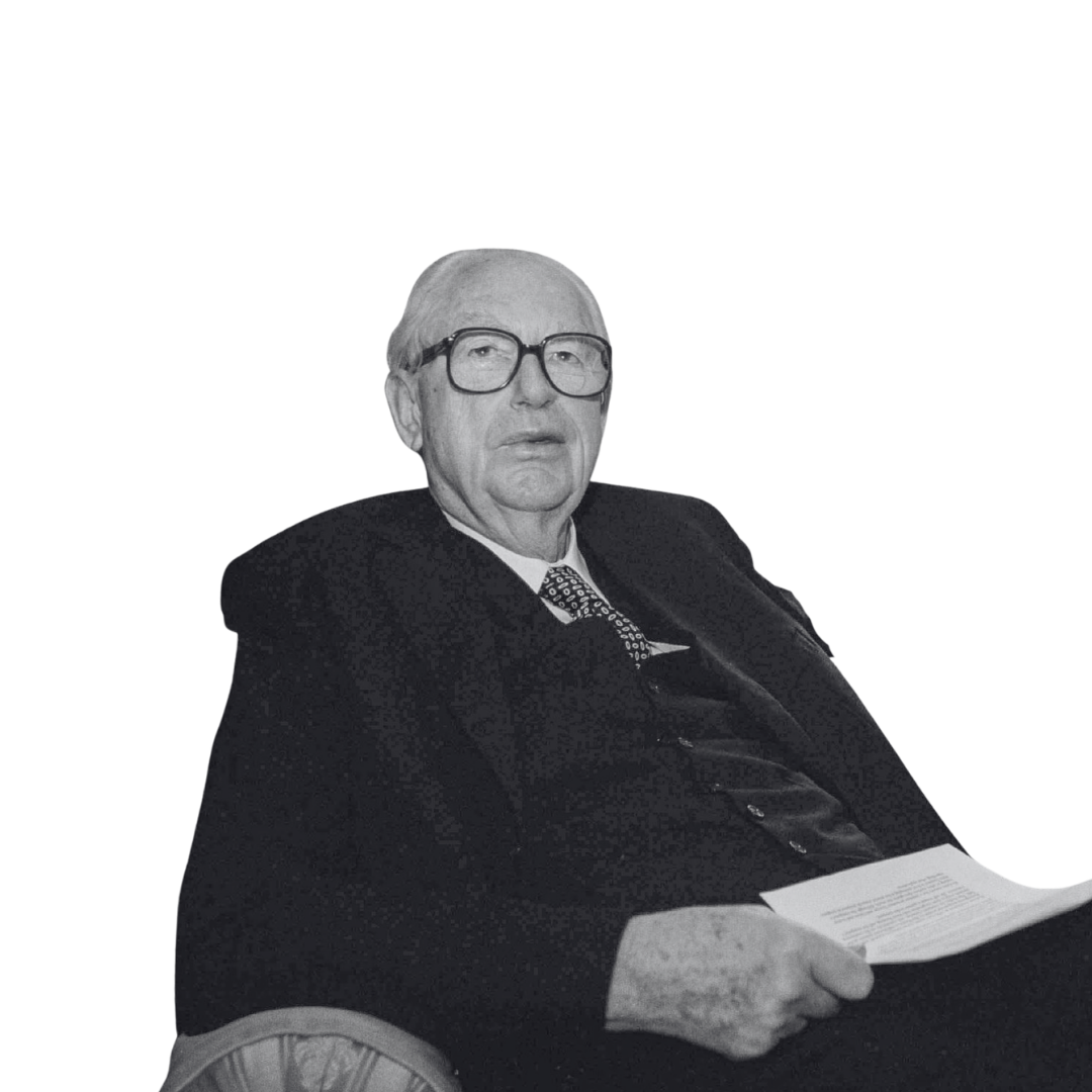An elderly man with glasses, wearing a suit and tie, sitting on a chair and holding a piece of paper.