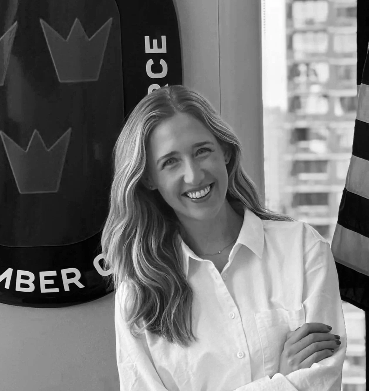 A smiling woman with long wavy hair outdoors near a body of water and trees.