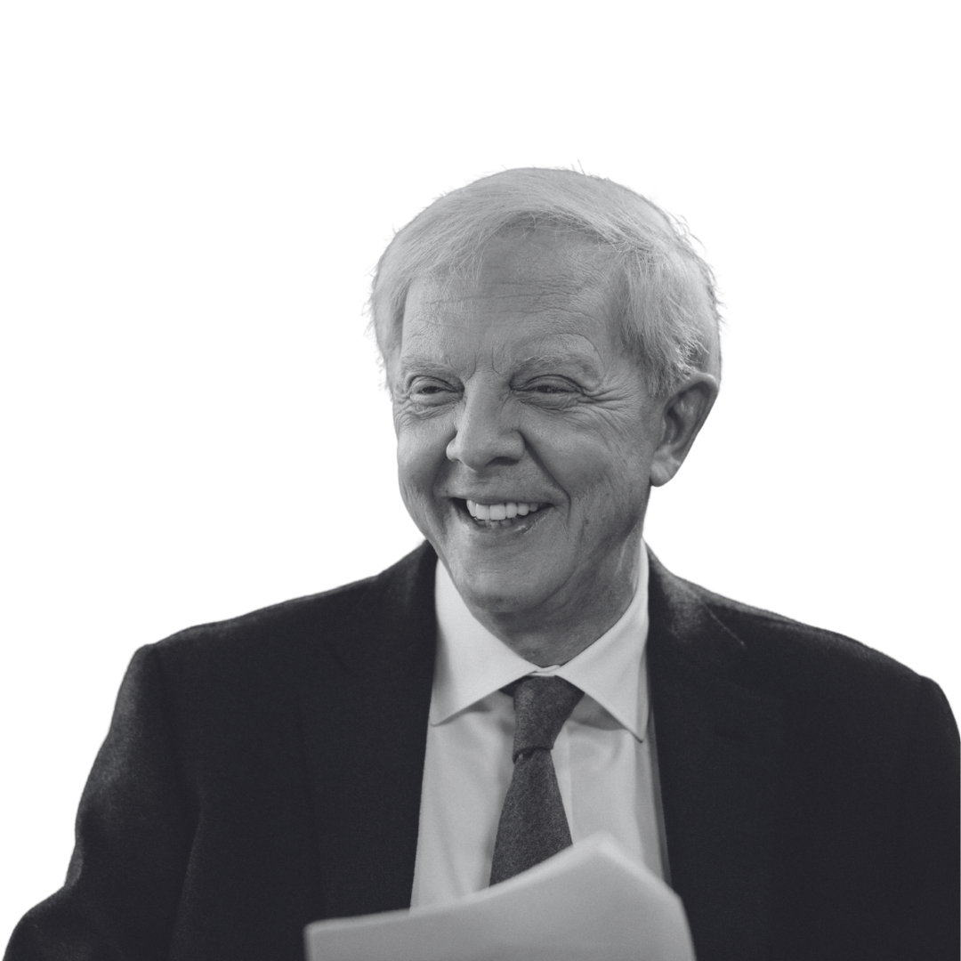 Black and white photo of an older man with light hair, smiling and wearing a suit and tie, holding a paper.