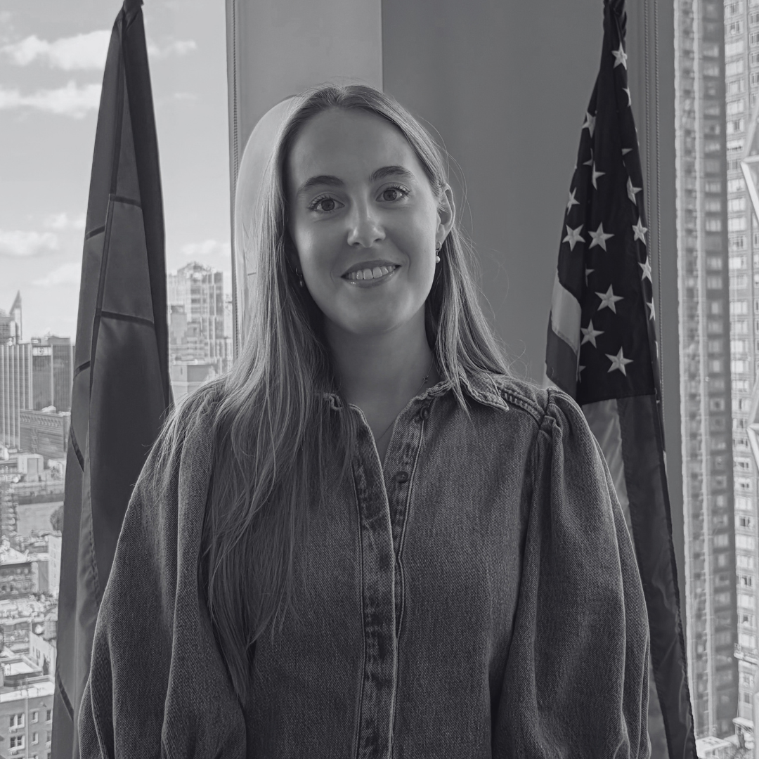 A woman standing in front of windows with city buildings outside, flanked by the United States flag and another flag.