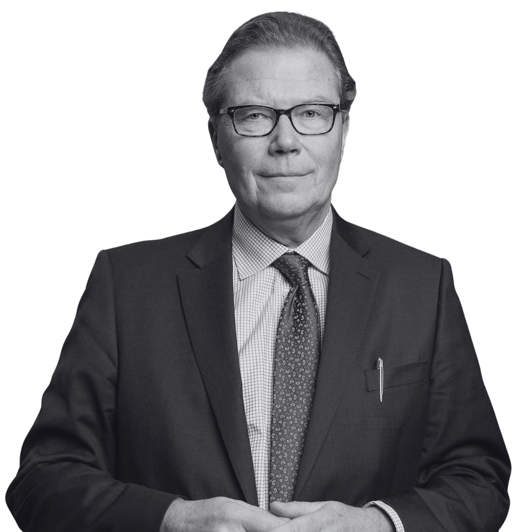Black and white portrait of an older man wearing glasses, a dark suit, a checkered shirt, and a patterned tie, standing with hands clasped in front.