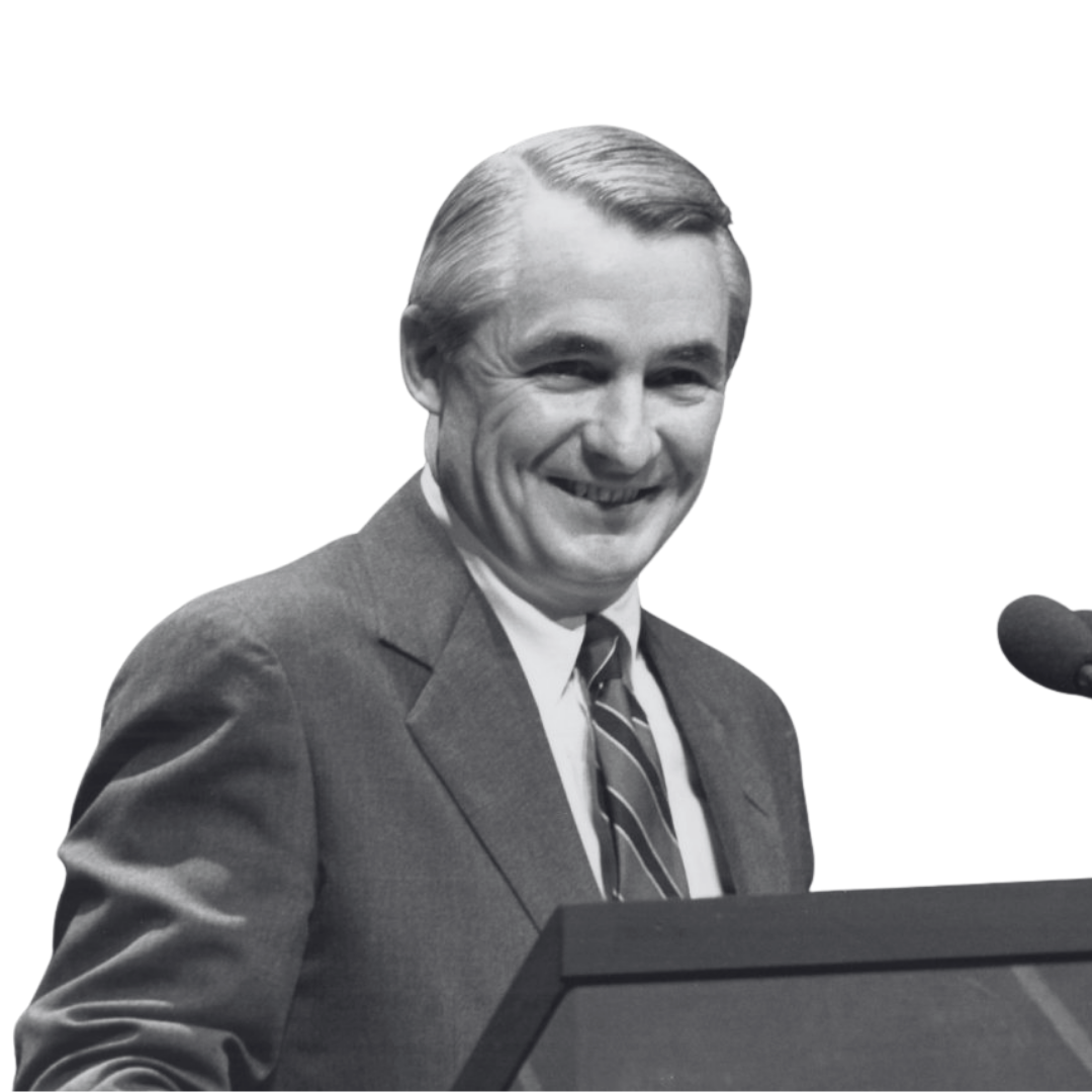Black and white photo of a smiling man in a suit and tie standing at a podium with a microphone.