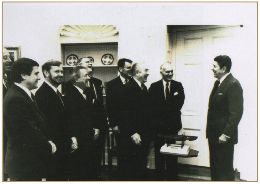 A black and white photo of a group of men in suits standing around President John F. Kennedy in the White House, having a conversation and smiling.