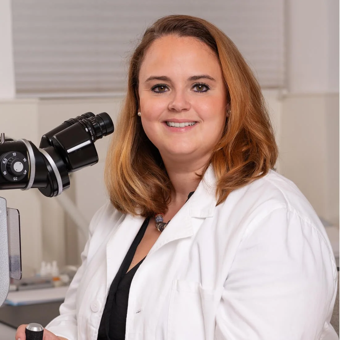 Dr. Annette Harrer-Seely, Kinderaugenärztin in 1130 Wien. Eine Ärztin in einem weißen Arztkittel steht neben einem Mikroskop in ihrem Praxisraum.