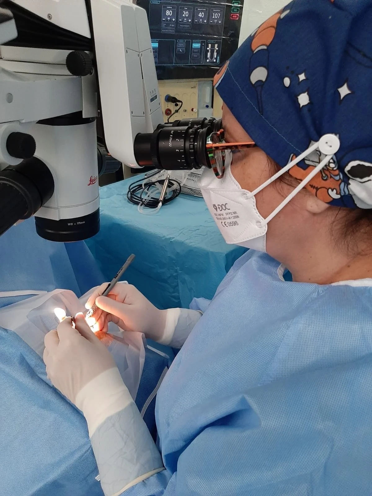 Ärzte im Operationssaal bei einer OP, der Arzt trägt eine chirurgische Maske und eine spezielle Kopfbedeckung, benutzt ein Mikroskop und arbeitet an einem kleinen Eingriff.