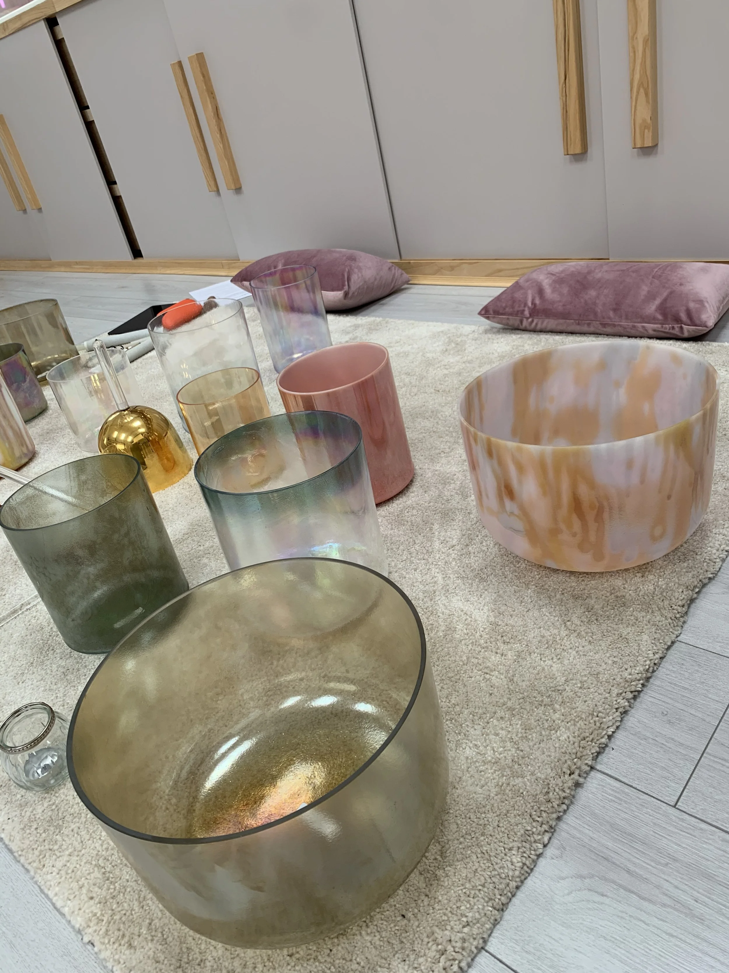 A collection of colorful glass singing bowls arranged on a carpeted floor, with soft cushions nearby.