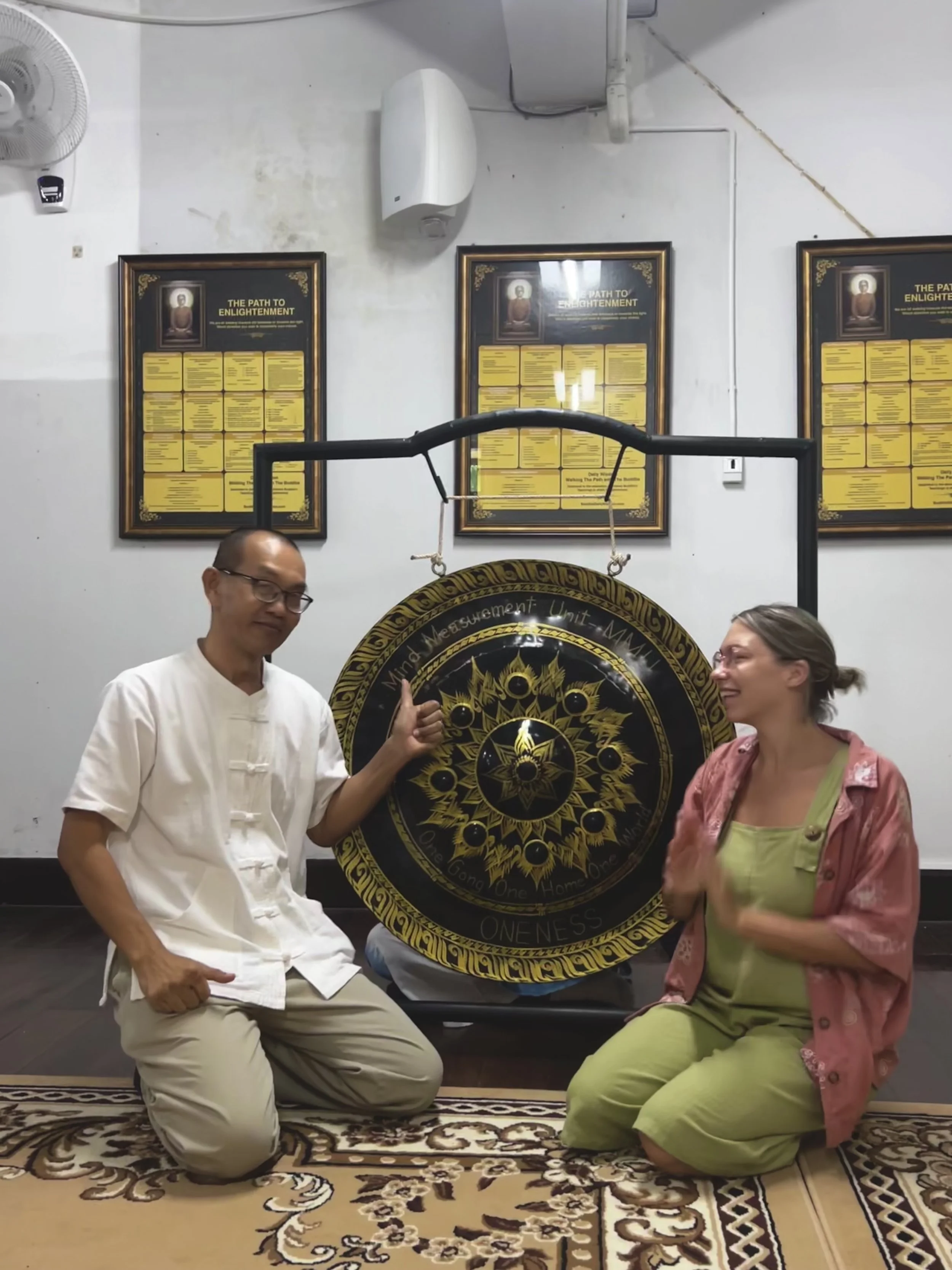 Two people kneeling in front of a large gong, with decorative patterns, inside a room with plaques on the wall. They are smiling and gesturing with enthusiasm, suggesting a joyful or celebratory moment.