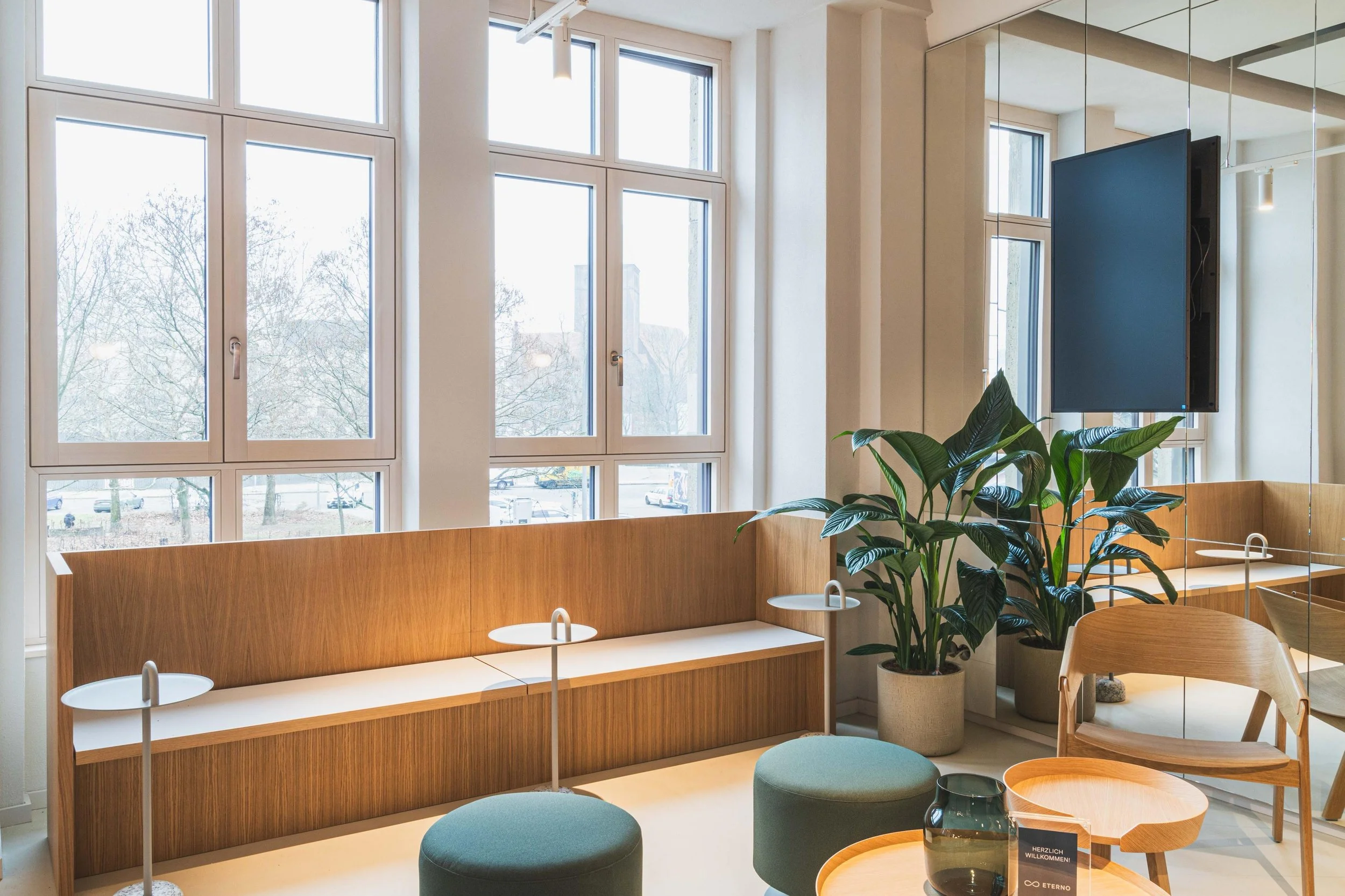 Moderner Lounge-Bereich mit großen Fenstern, Grünpflanzen, Sitzgelegenheiten aus Holz, einem an der Wand montierten Fernseher und modernen Möbeln.