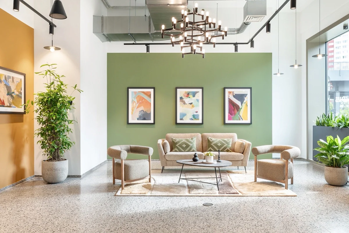Modern hotel lobby with a beige sofa, two matching armchairs, and a coffee table on a patterned rug. The background features a green wall with three framed abstract artworks, large windows to the right, and potted plants on each side.