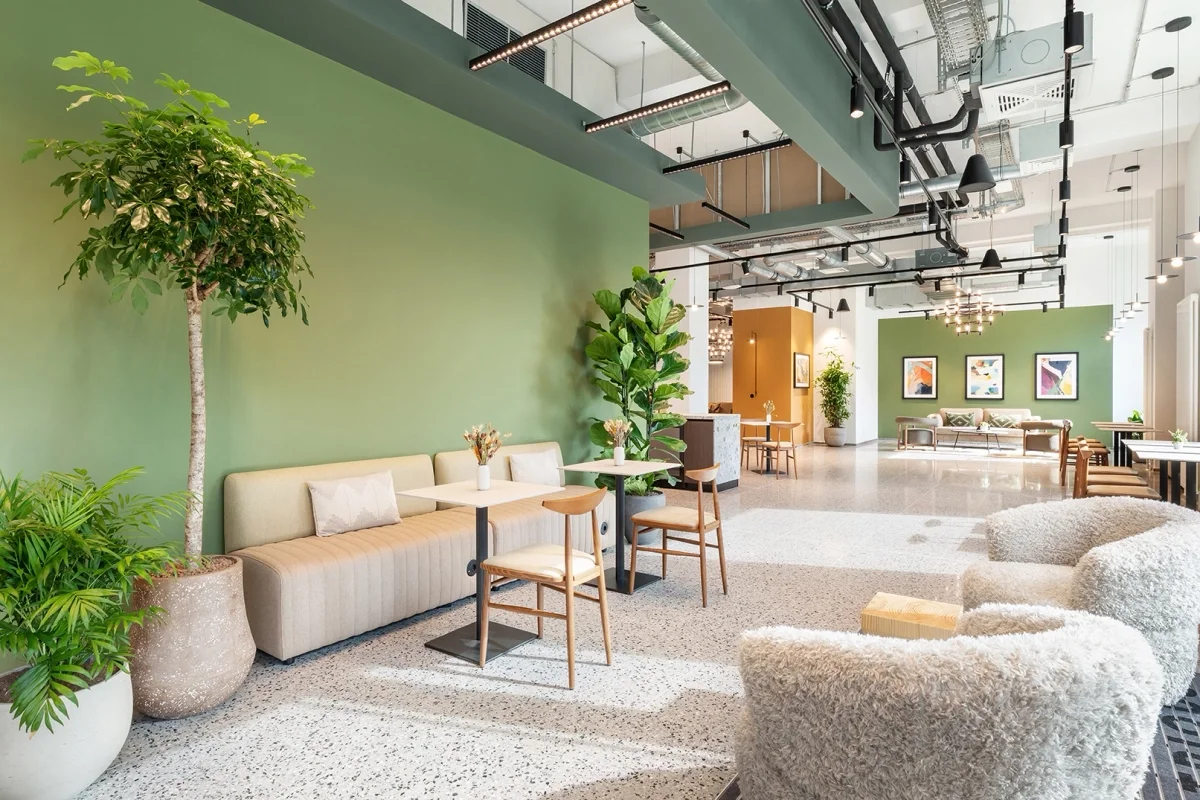 Bright, modern café interior with green accent walls, various green plants, cozy seating including plush chairs and a sofa, small tables with vases, and contemporary art on the walls.