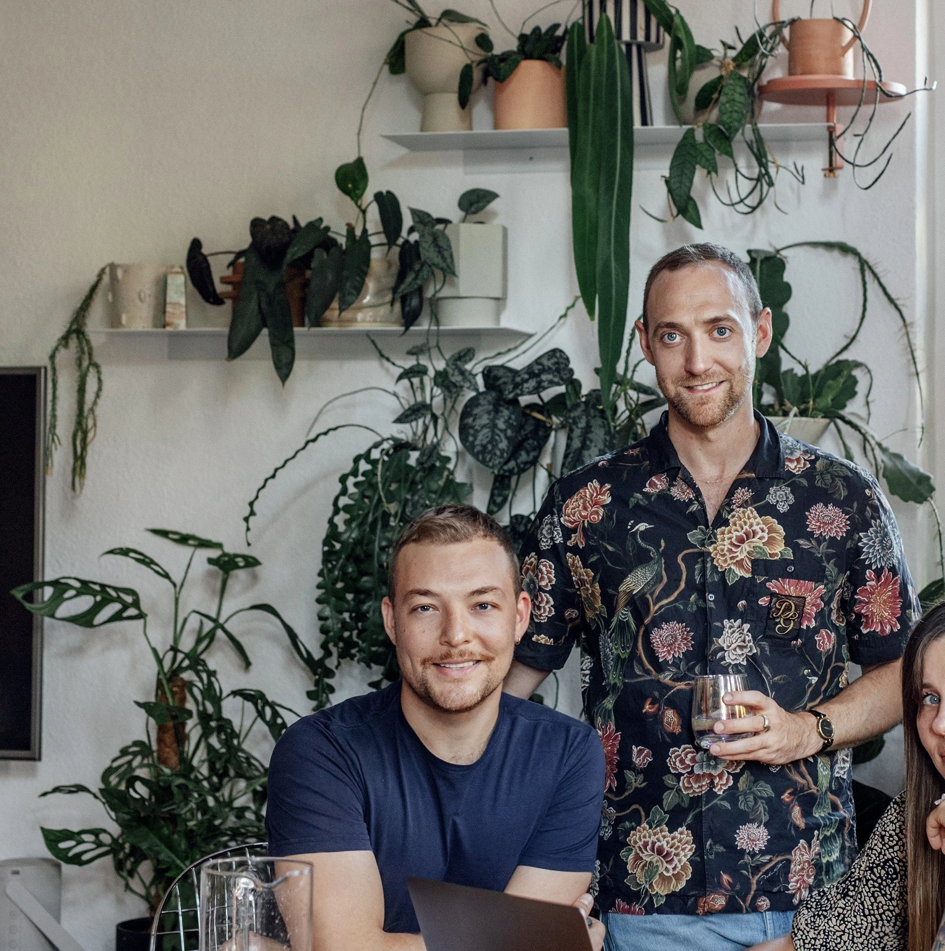Two men in a room filled with various houseplants. One is seated with a laptop, wearing a blue shirt, and the other is standing beside him holding a glass, wearing a floral shirt. A woman is partially visible on the right.