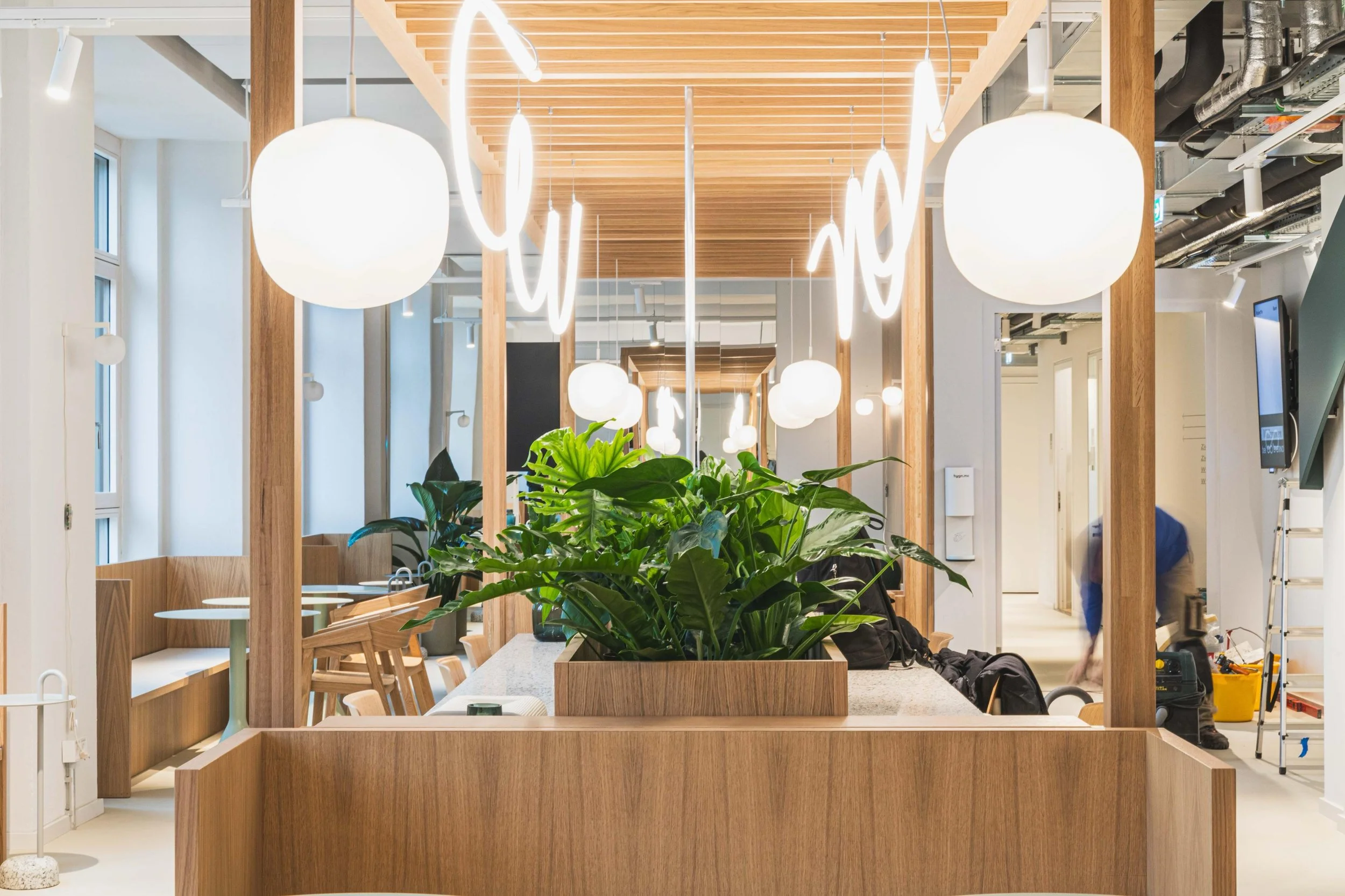 Innenraum eines modernen Büros mit Holzakzenten, hängenden weißen Pendelleuchten und Grünpflanzen in der Mitte. Entlang der Fenster befinden sich Sitzbereiche mit Stühlen und Tischen, und im Hintergrund sind einige Geräte und eine arbeitende Person zu sehen.