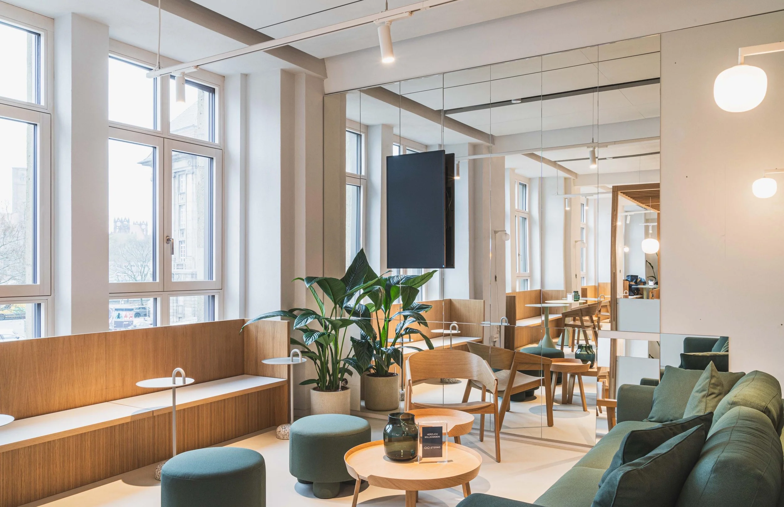 Moderner Aufenthaltsbereich mit großen Fenstern, grünem Sofa, Holzstühlen und Pflanzen.