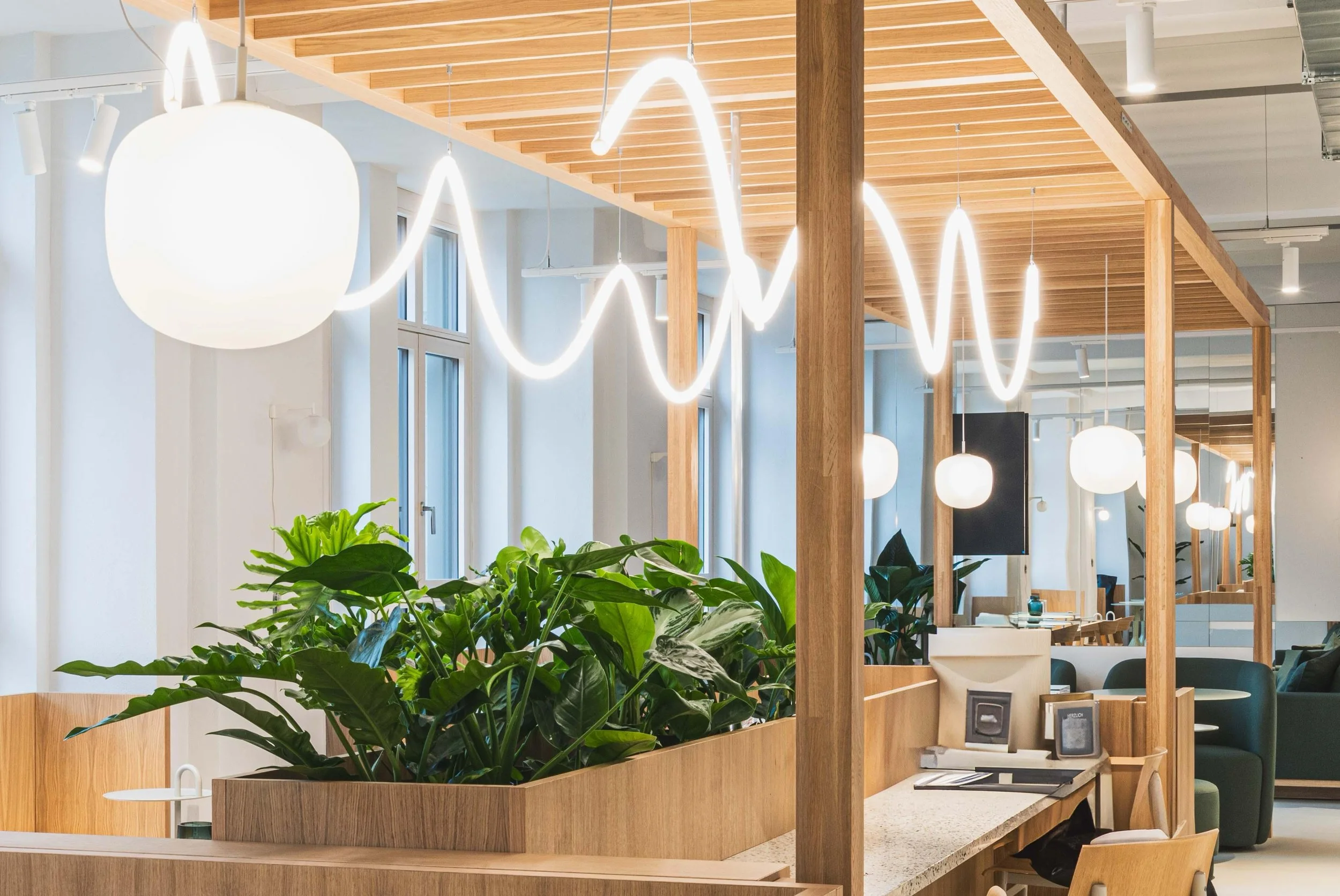 Modernes Bürointerieur mit hölzernen Trennwänden, Grünpflanzen, hängenden weißen Kugelleuchten und einer dekorativen kurvigen Neonleuchte.