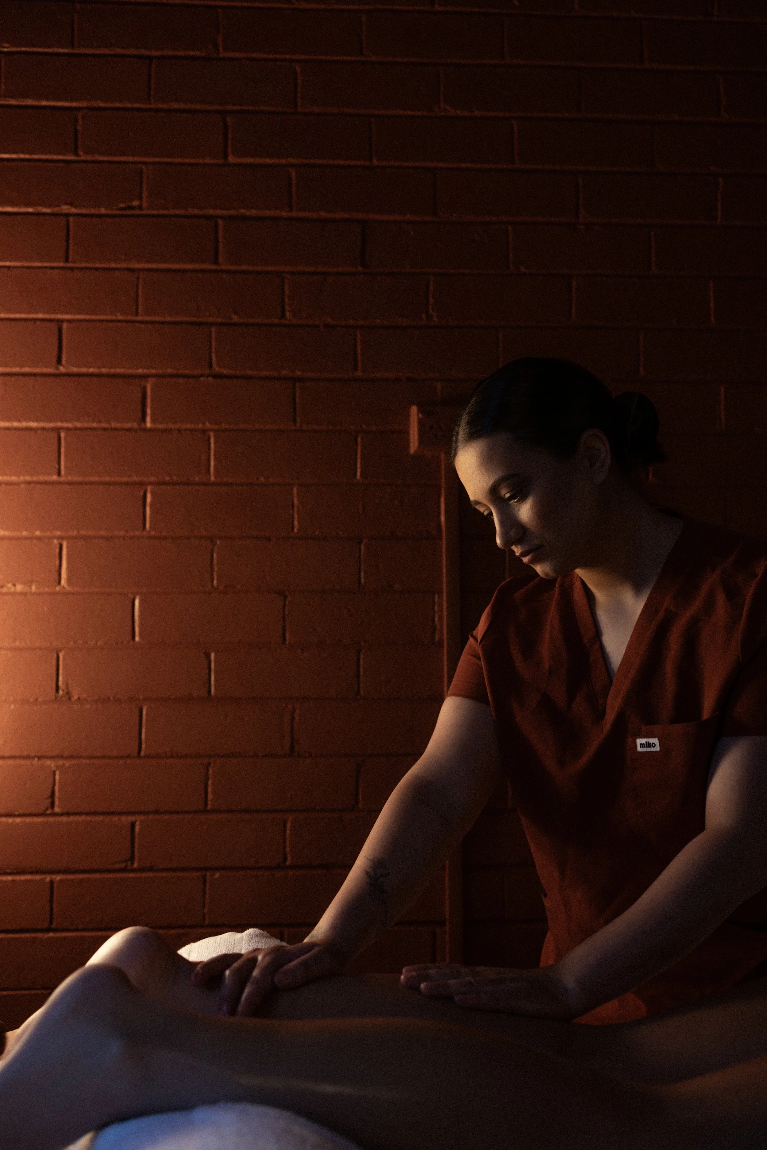 A massage therapist giving a massage in a dimly lit room with a brick wall in the background.