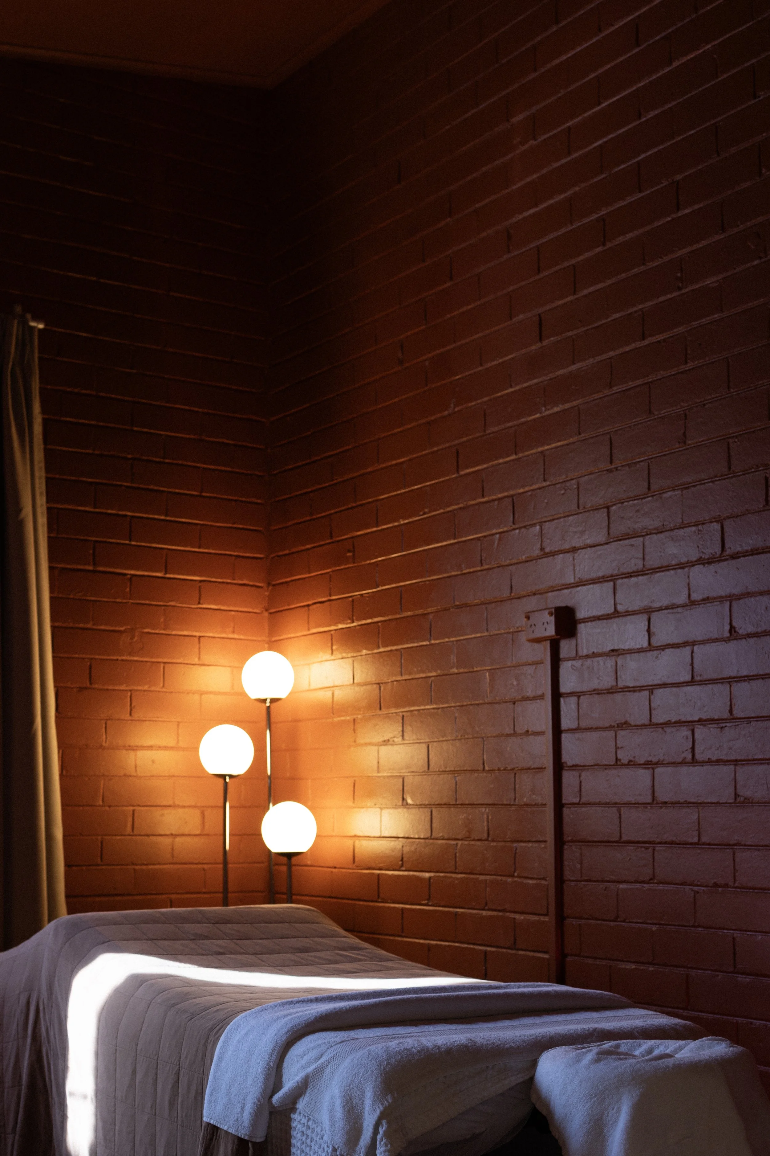A bedroom with a brick wall, a bed with a beige quilt, and a floor lamp with three globes emitting warm light.