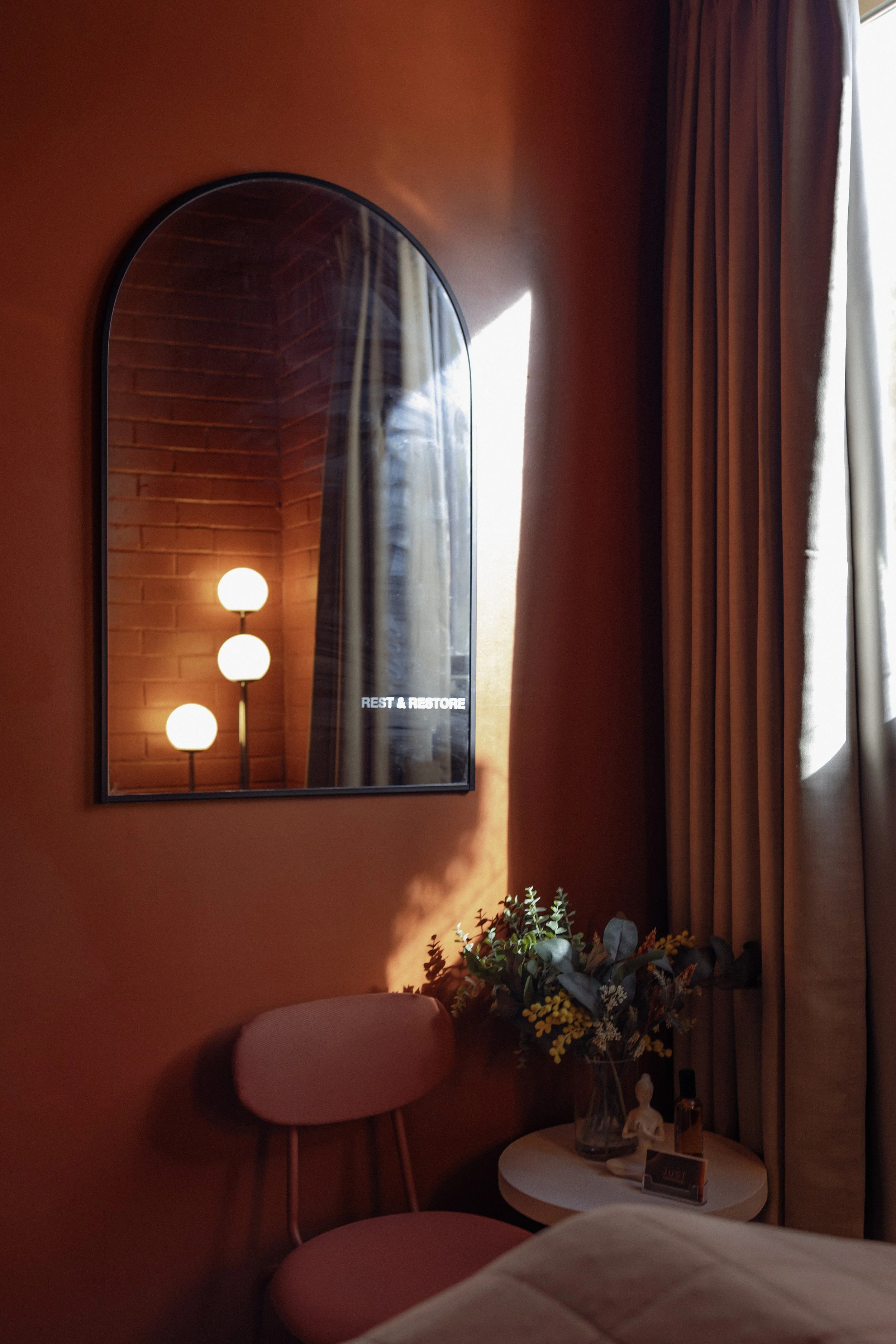 A cozy corner of a room with an orange wall, a mirror reflecting a brick wall, and a lamp with three round bulbs. There is a pink chair and a small table with a vase of flowers, a small statue, and a perfume bottle near a window with cream curtains.