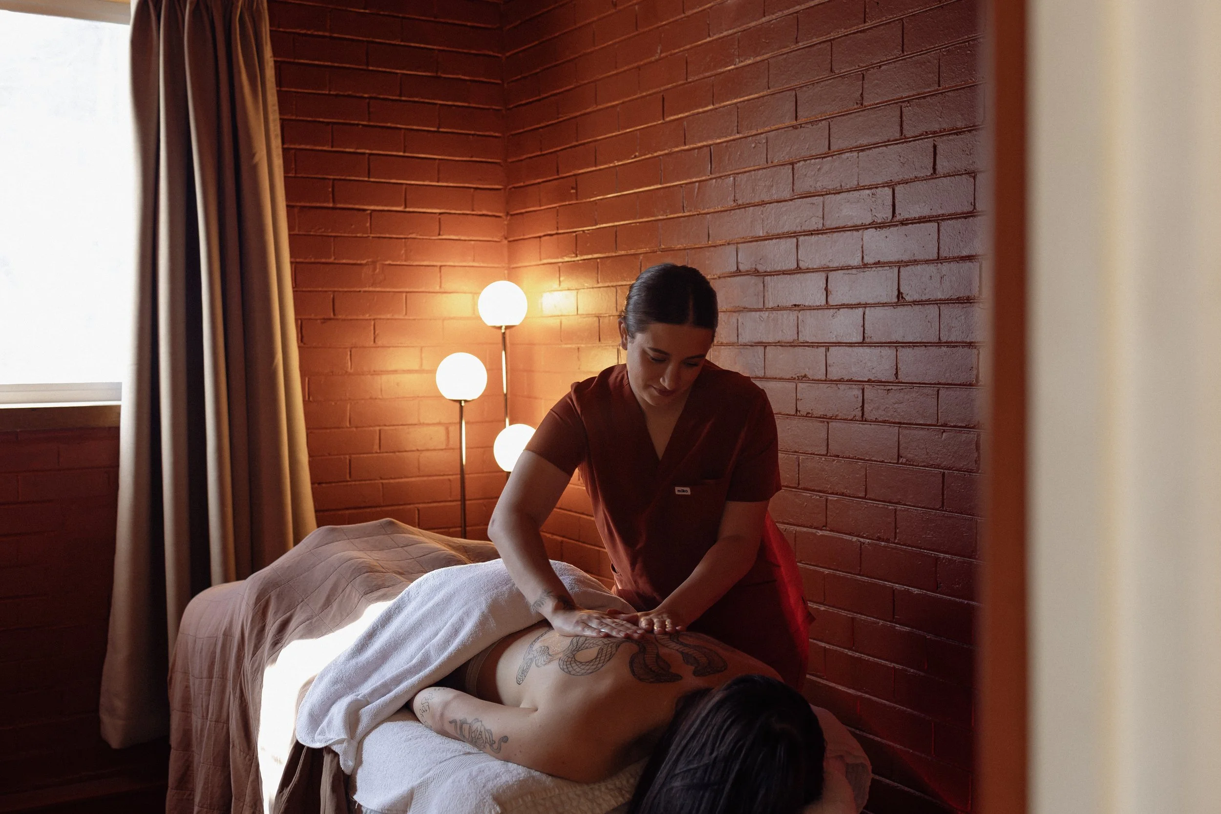 A woman giving a massage to a person lying face-down on a massage table in a dimly lit room with a brick wall and a floor lamp.
