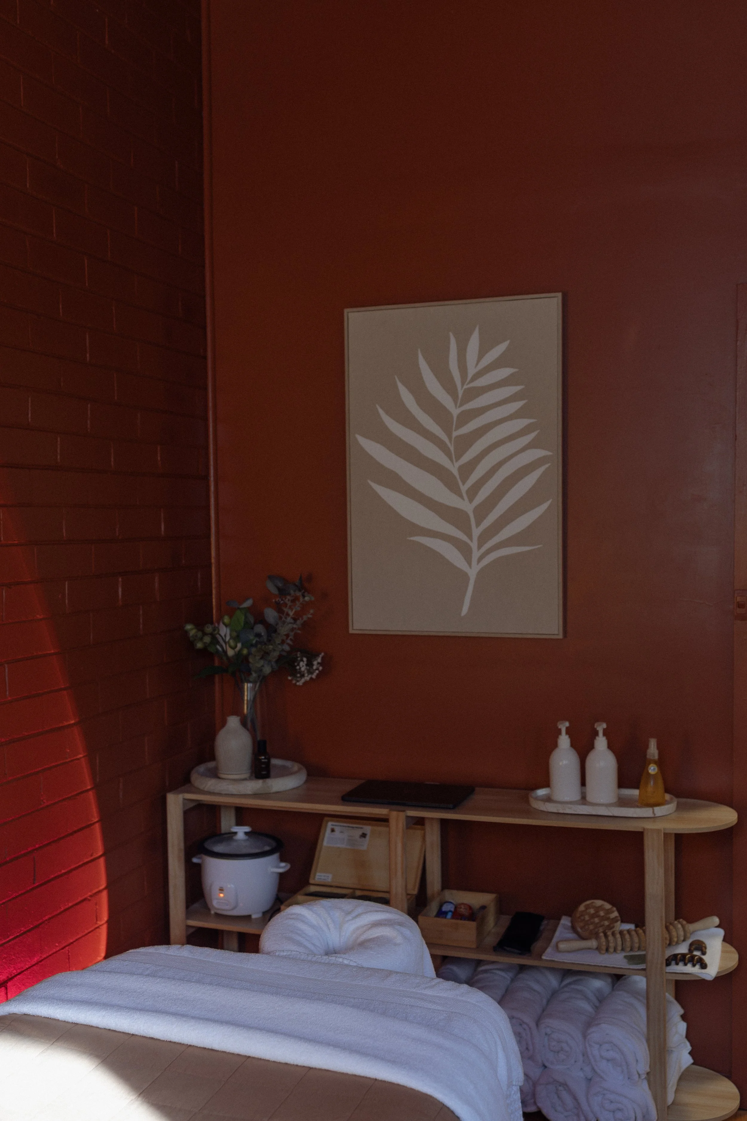 A massage therapy room featuring a bed with a white rolled towel, a wooden shelf with bottles, a rice cooker, and framed wall art of a large leaf design, all against earthy red walls decorated with a botanical print.