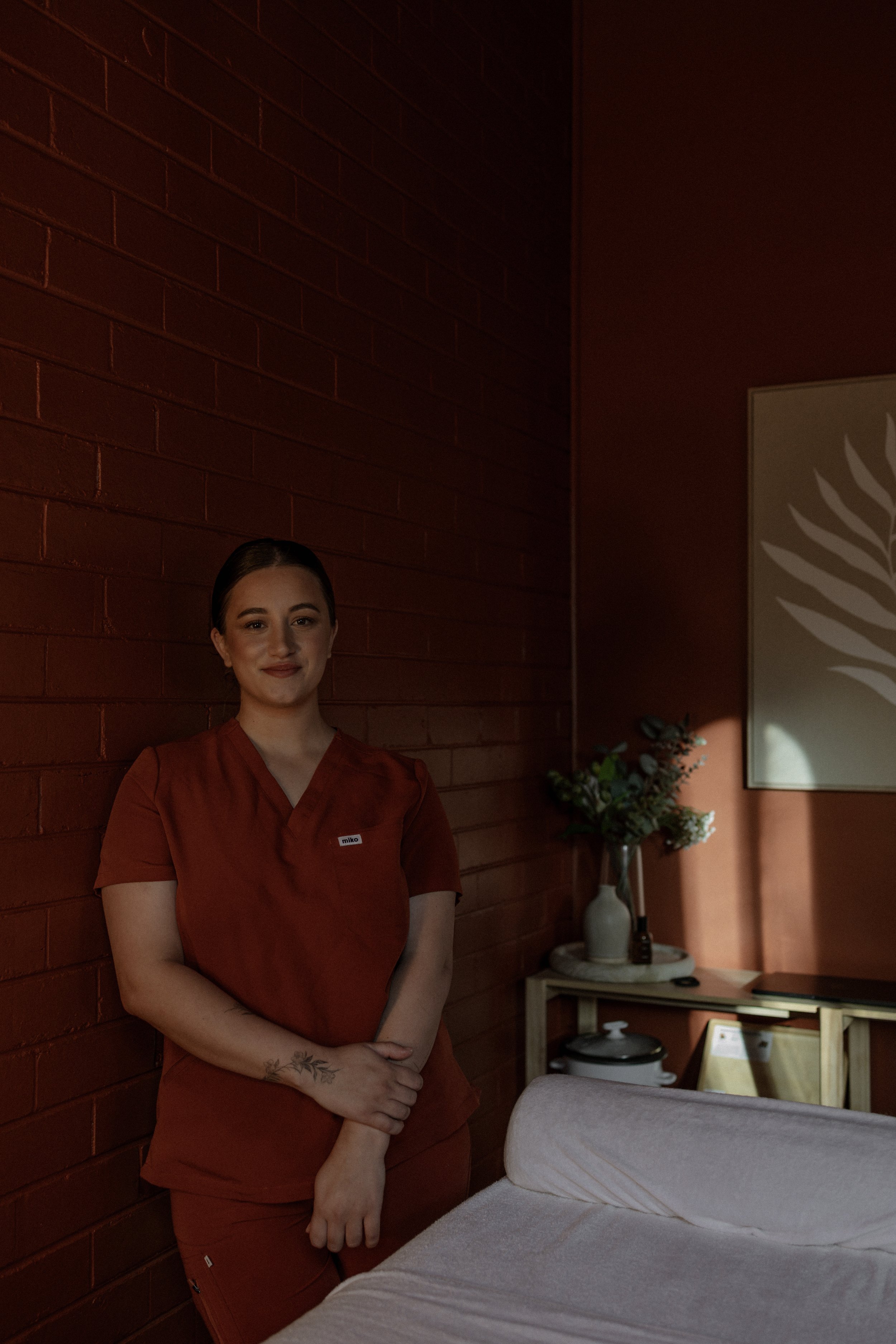 A young woman standing in a dimly lit room with a brick wall, wearing red scrubs, smiling, with a tattoo on her wrist, beside a bed and a small table with a vase of flowers and some objects.