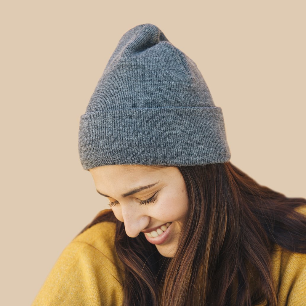 Smiling person wearing a gray knit beanie and a yellow sweater on a neutral background.