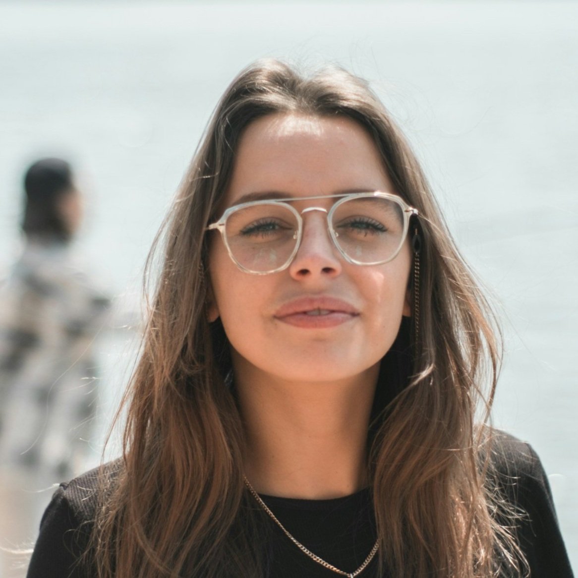 Young woman with glasses standing outdoors, wearing a black shirt, with a blurred person and water in the background.