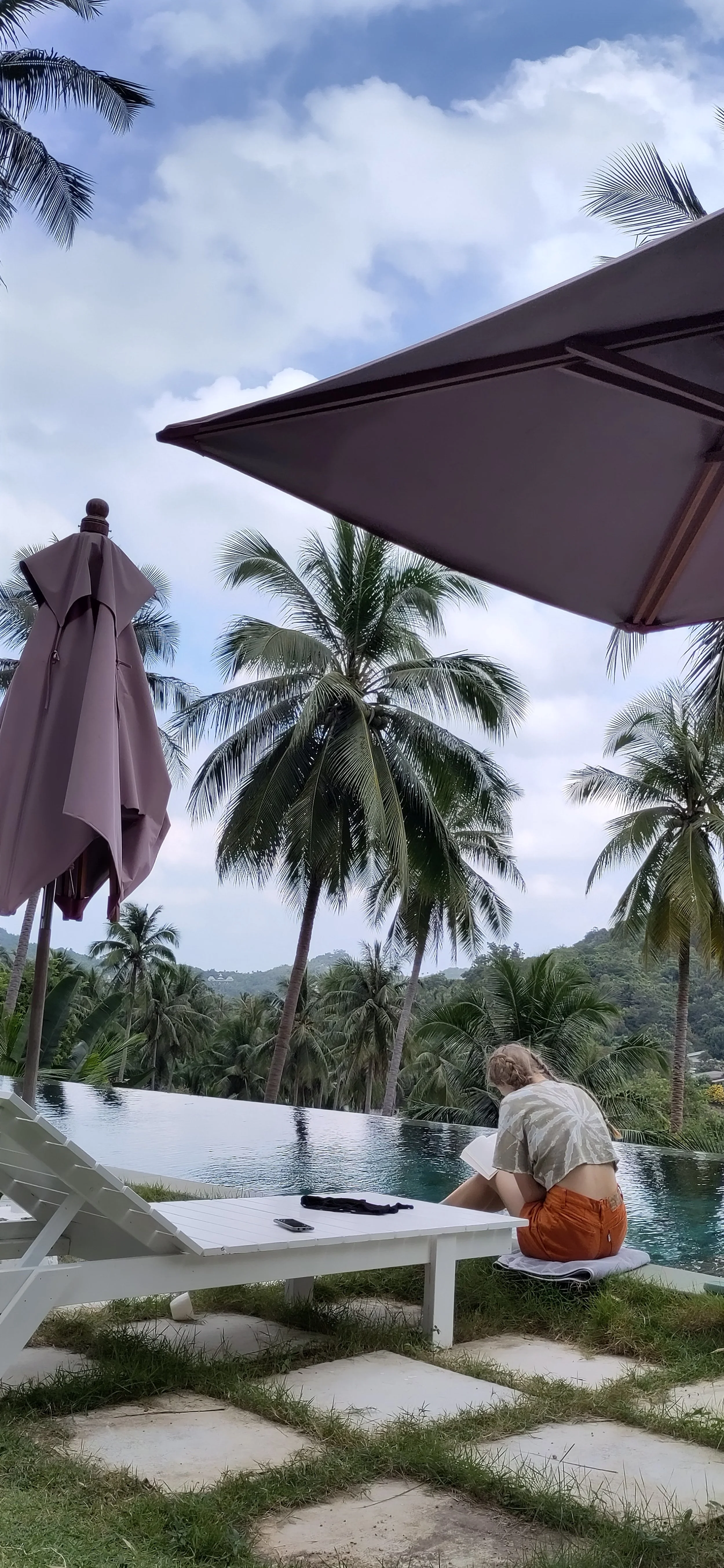 Eine Frau liest an einem Pool mit Palmen im Hintergrund, umgeben von Liegestühlen und Sonnenunterschirm. Herrlicher Blick auf eine bewaldete Hügellandschaft.