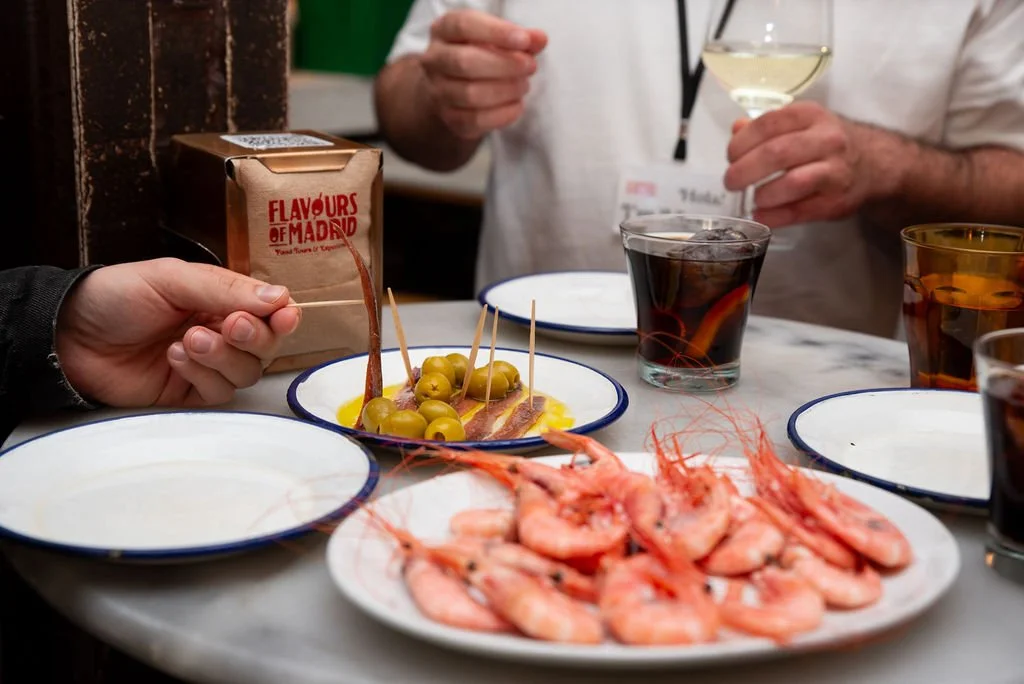 A table with a plate of cooked shrimp, a dish of green olives with toothpicks, and glasses of white wine, red wine, and soda, with people serving drinks in the background.