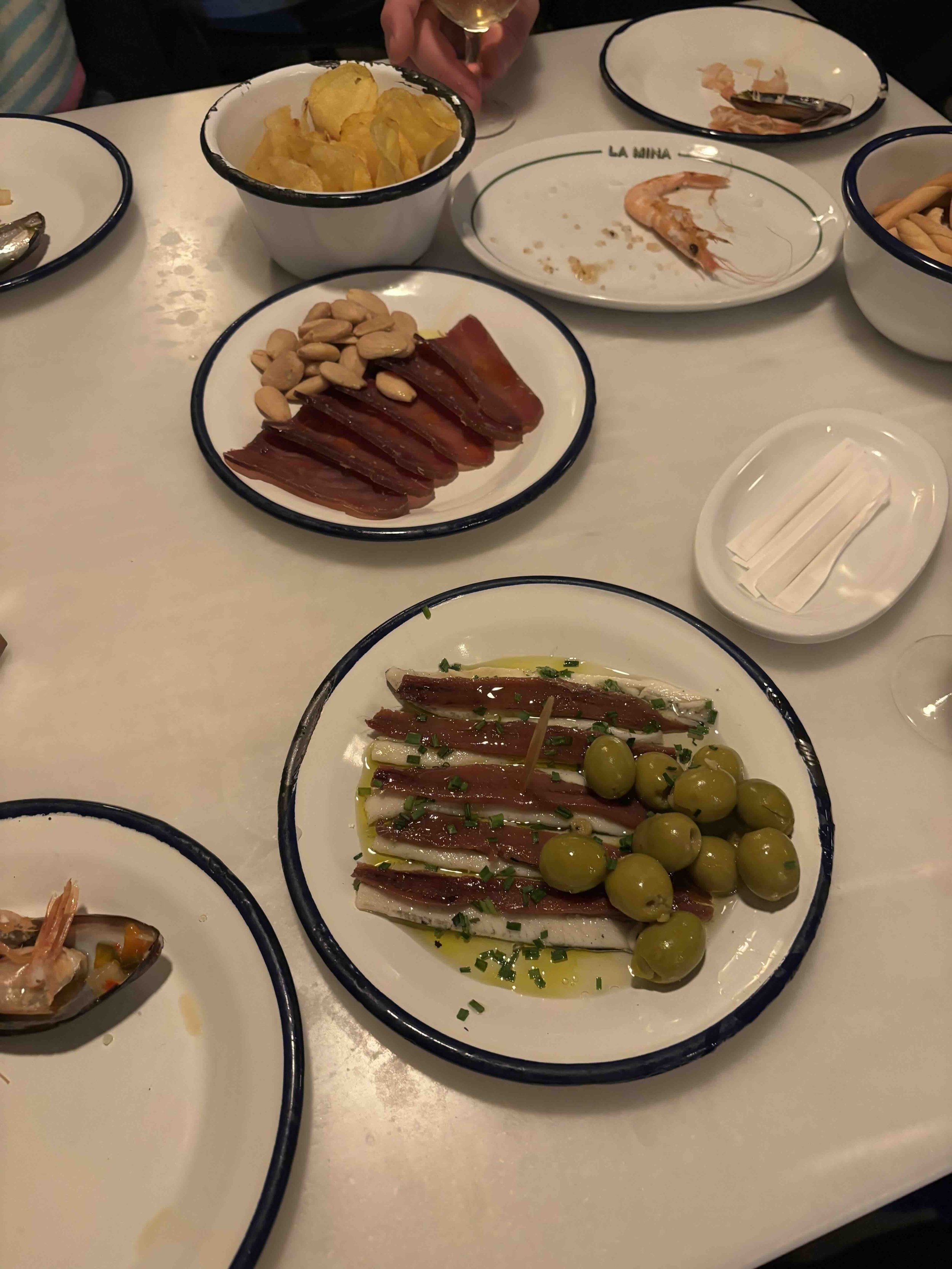 Empty plates with seafood and snacks on a white table, including a plate with anchovies, green olives, and chopped herbs, another with sliced cured meat and almonds, and bowls with chips and other appetizers.