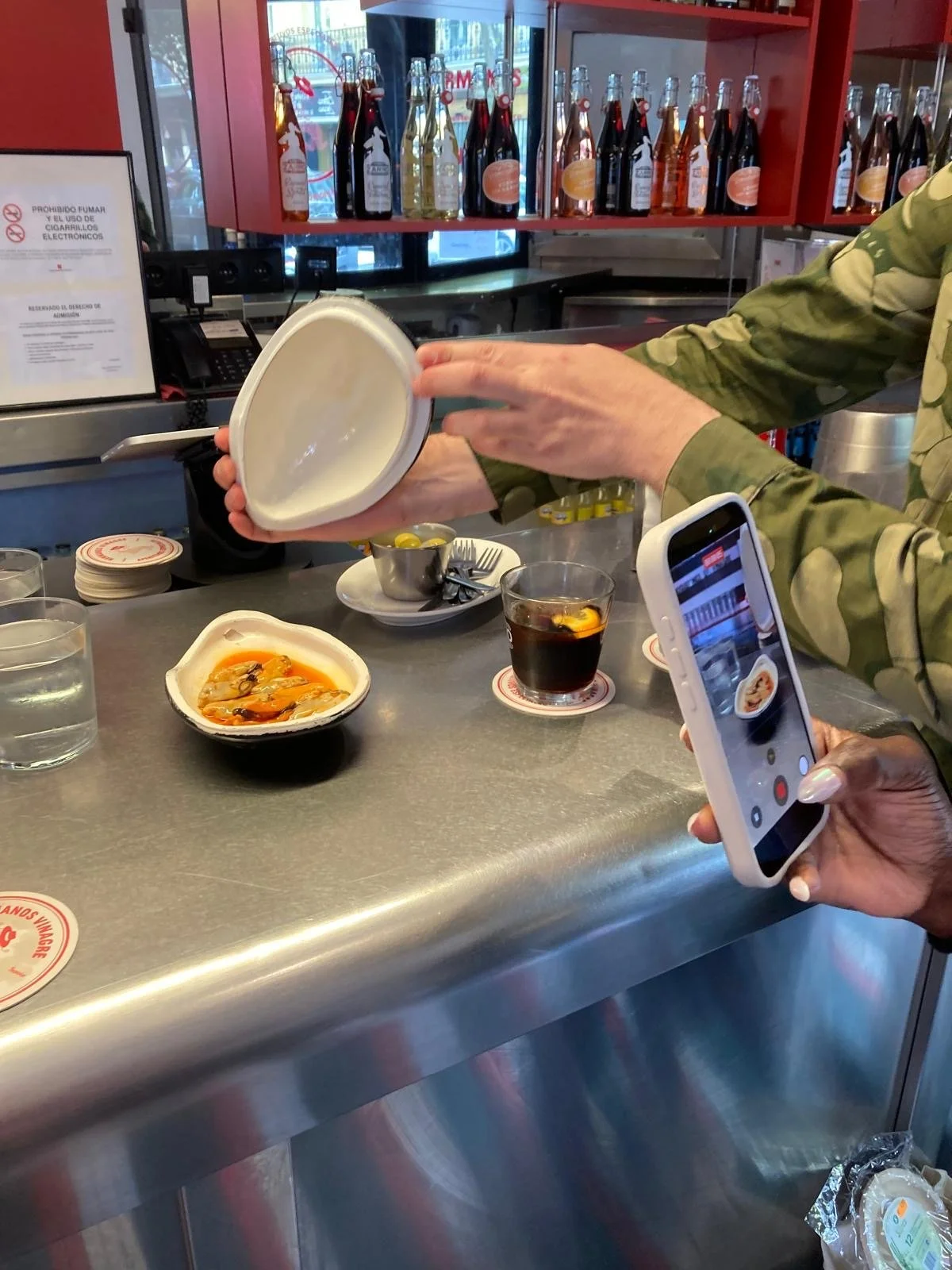 Person taking a photo of a bowl with seafood and sauce at a restaurant counter, with drinks and condiments nearby.