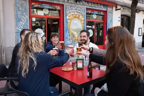 A group of six friends enjoying drinks and toasting at an outdoor table outside a bar or restaurant.
