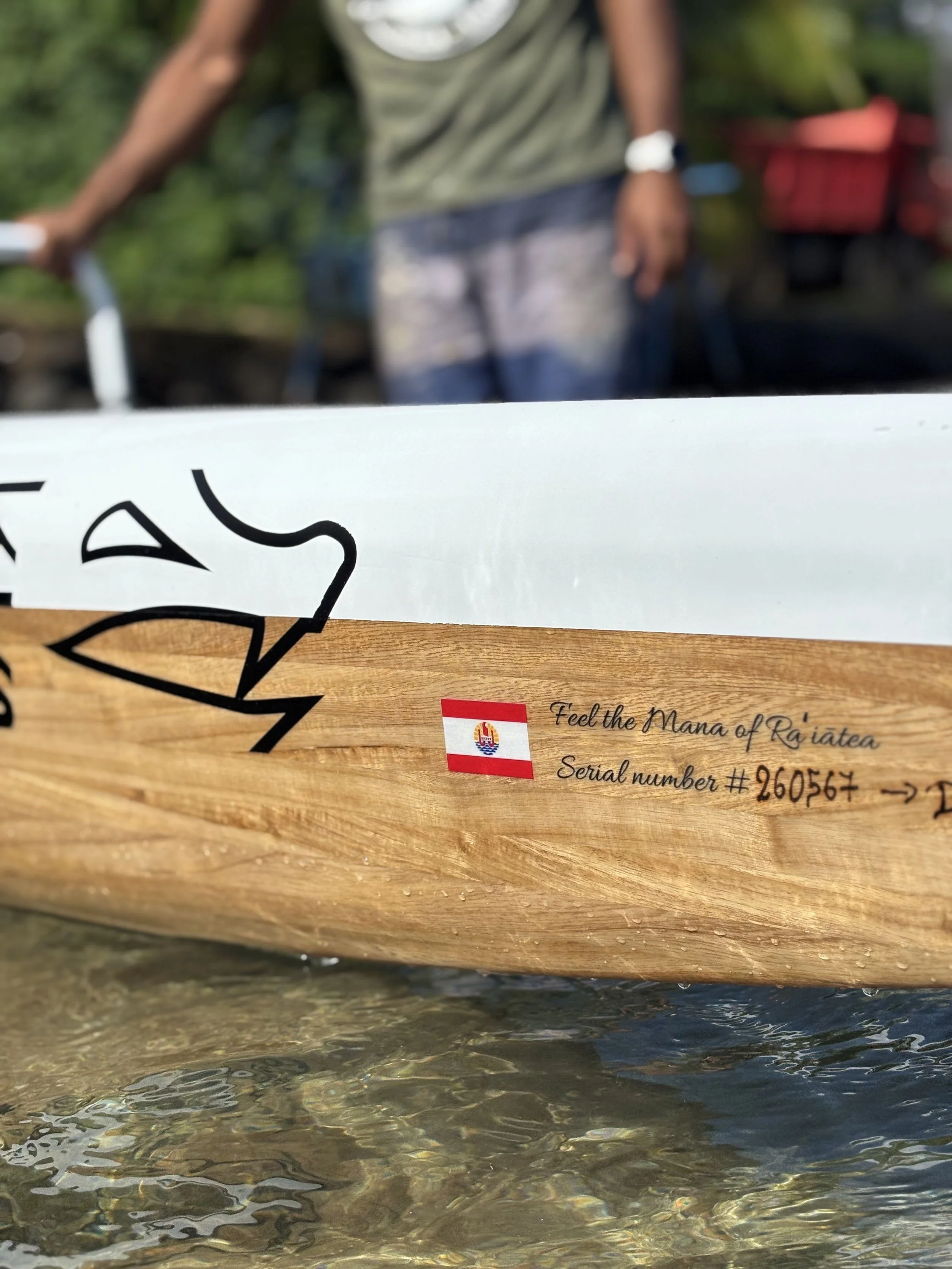 Un bateau en bois flotte dans une rivière, avec une étiquette indiquant un numéro de série et une phrase en anglais « Feel the Mana of Rájatea ». Retranscription du texte : 'Serial number #260567'.