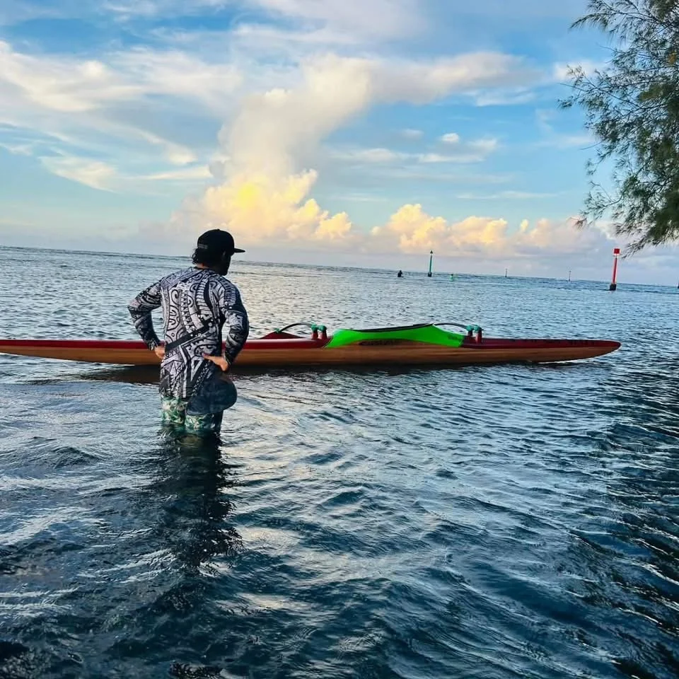 Une personne debout dans l'eau de mer, portant un chapeau noir et un vêtement à motifs, face à une pirogue tahitienne ou canoe coloré, dans un paysage marin avec un ciel partiellement nuageux.