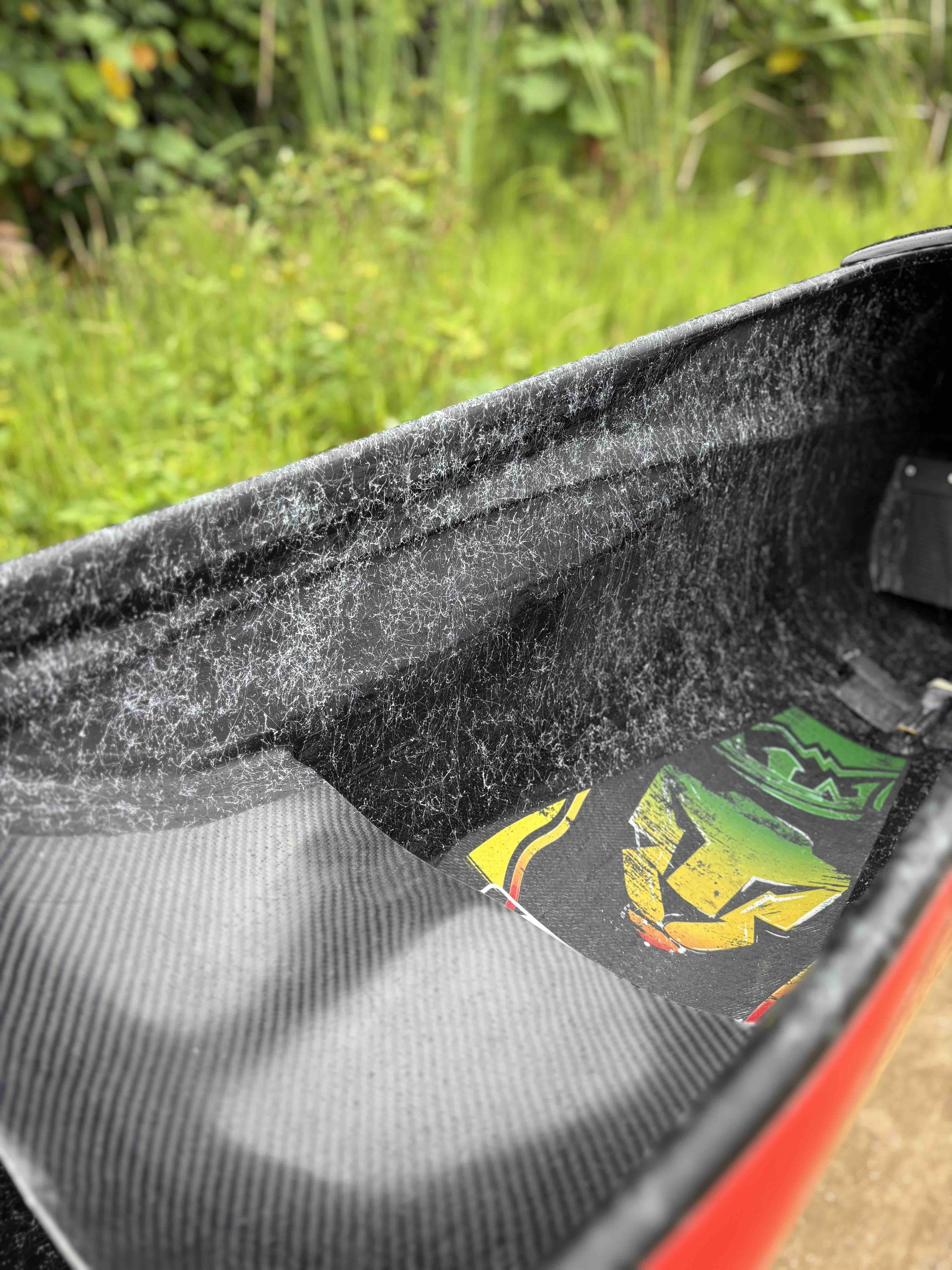 Une partie d'un vélo ou d'une planche de skateboard, avec une surface en fibre de carbone noire et une étiquette colorée, repose sur le sol dans un environnement extérieur verdoyant.