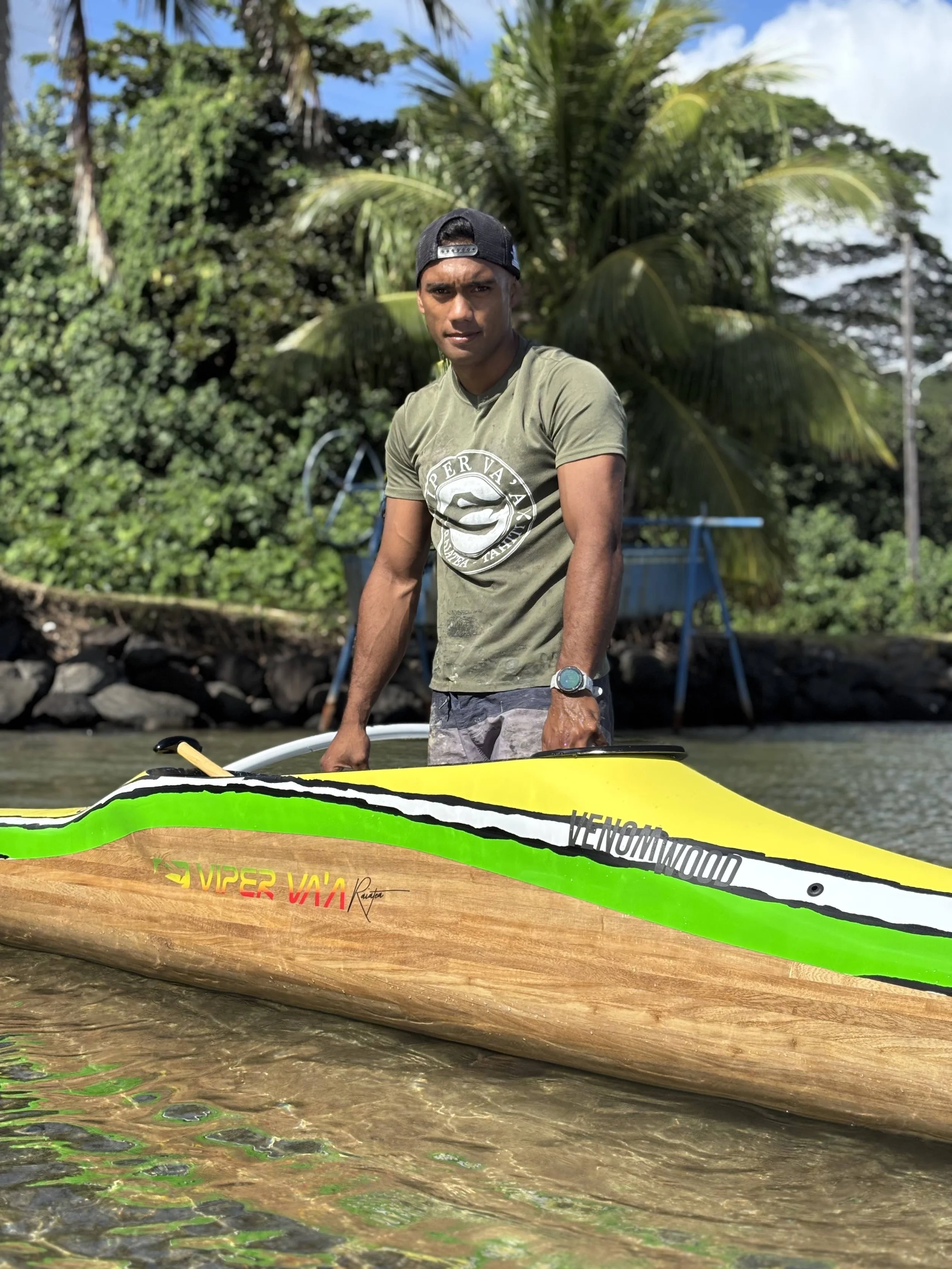 Jeune homme debout près d'une pirogue va'a en bois et en fibre de verre, portant un t-shirt vert, un casquette de baseball noir et une montre, dans une zone tropicale avec des palmiers et un ciel bleu, probablement à la plage.