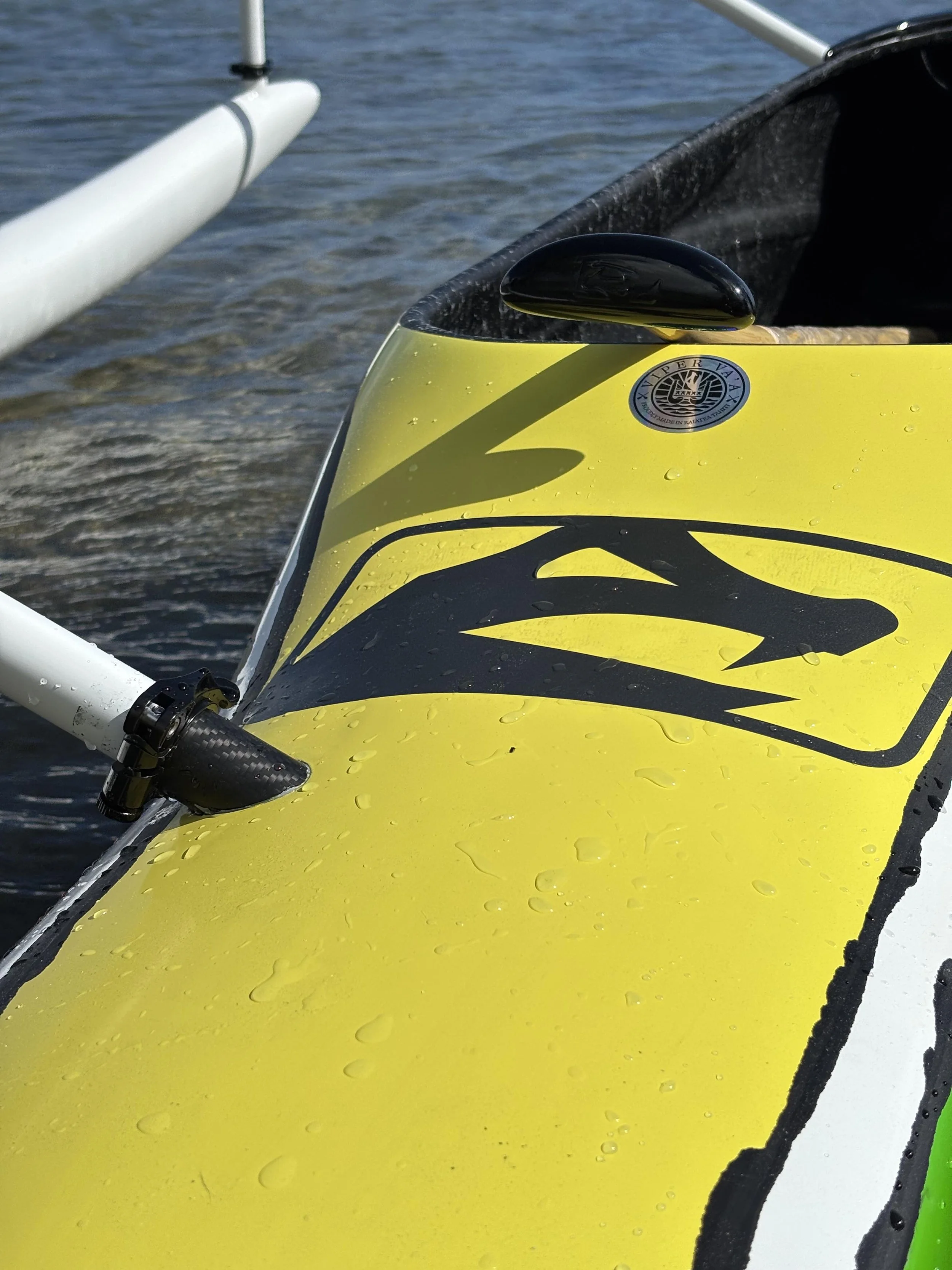 Vue rapprochée d'une section de va'a ou de pédalier avec un pont, un cockpit noir, un support noir, et une partie du flotteur en arrière plan, avec de petites gouttelettes d'eau sur la surface.