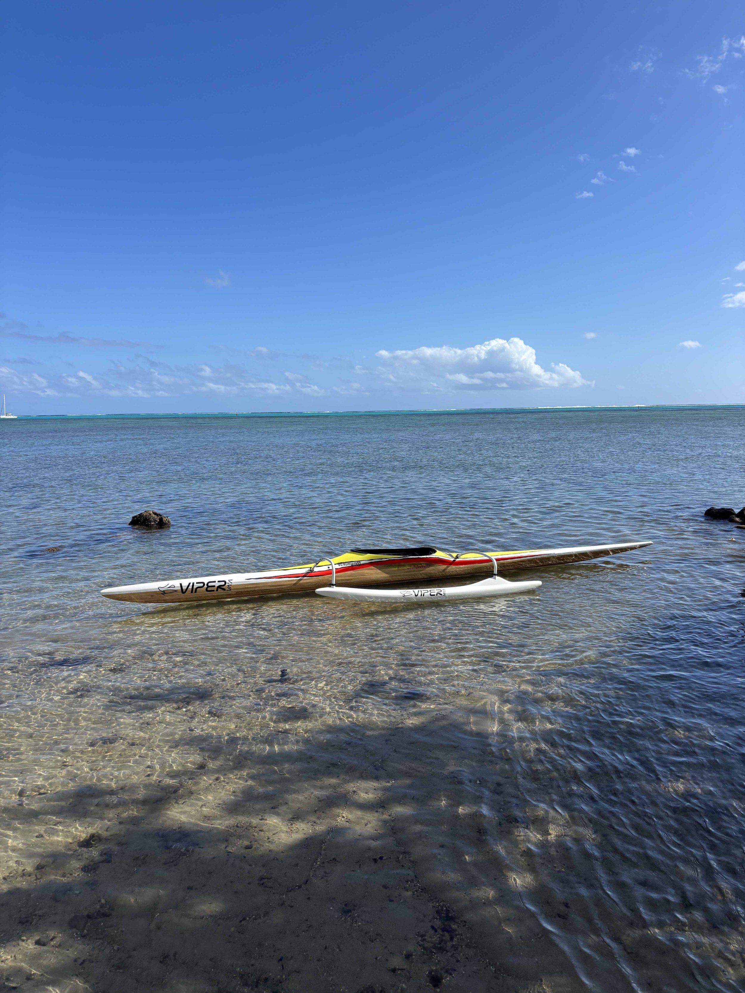 pirogue tahitienne blanche et jaune avec le mot 'VIPER' posé sur la plage au bord de l'eau, avec un ciel bleu et quelques nuages dans l'horizon.