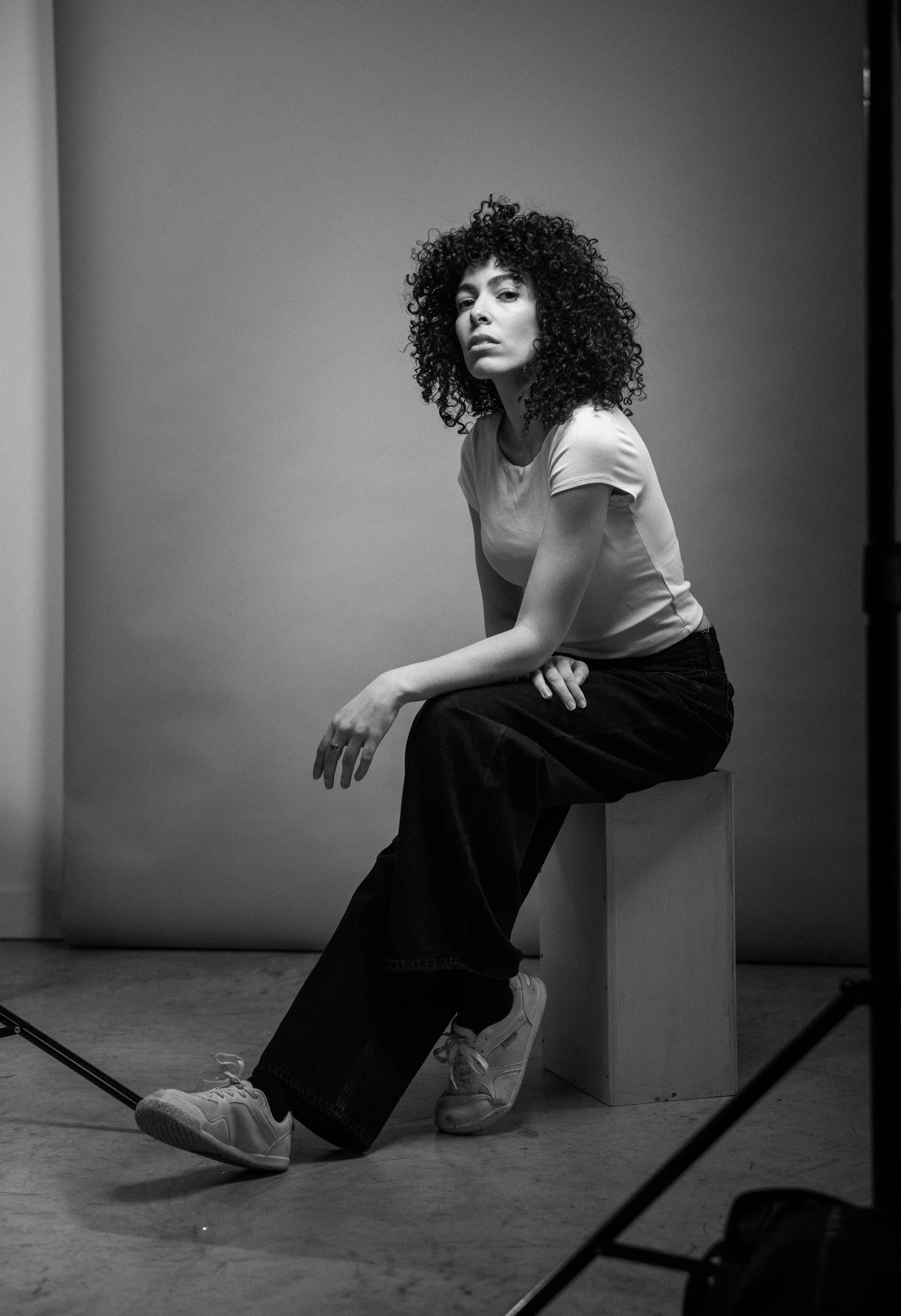 Une femme aux cheveux bouclés, assise sur un cube, posant pour une séance photo en noir et blanc.