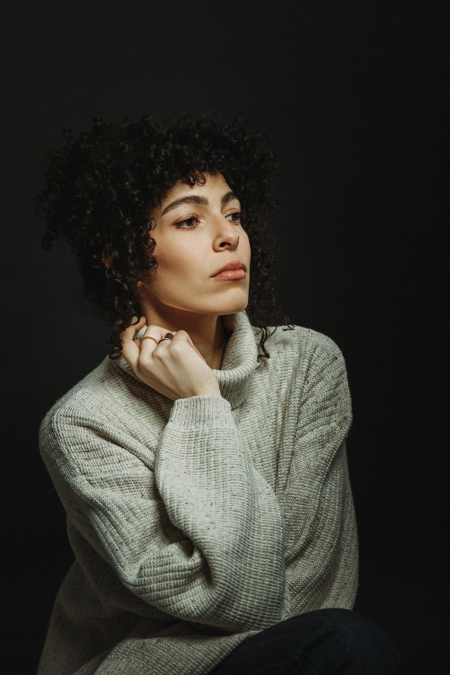 Portrait d'une femme avec des cheveux bouclés, portant un pull gris clair, assise contre un fond noir, regardant pensivement.
